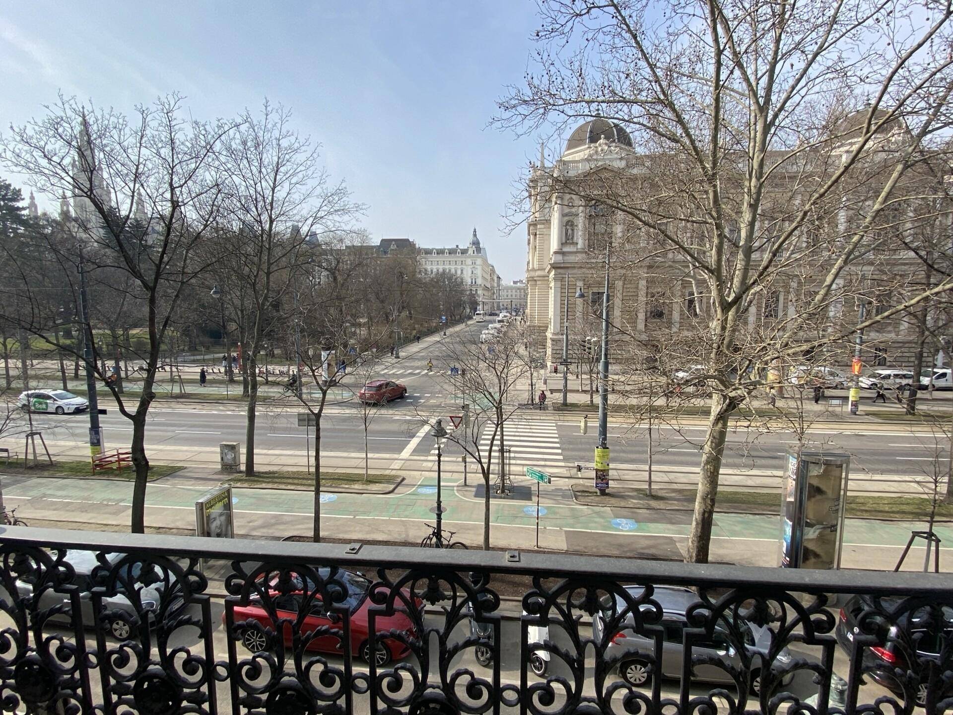 Blick vom Balkon auf eine belebte Stadtstraße mit Bäumen und historischen Gebäuden.