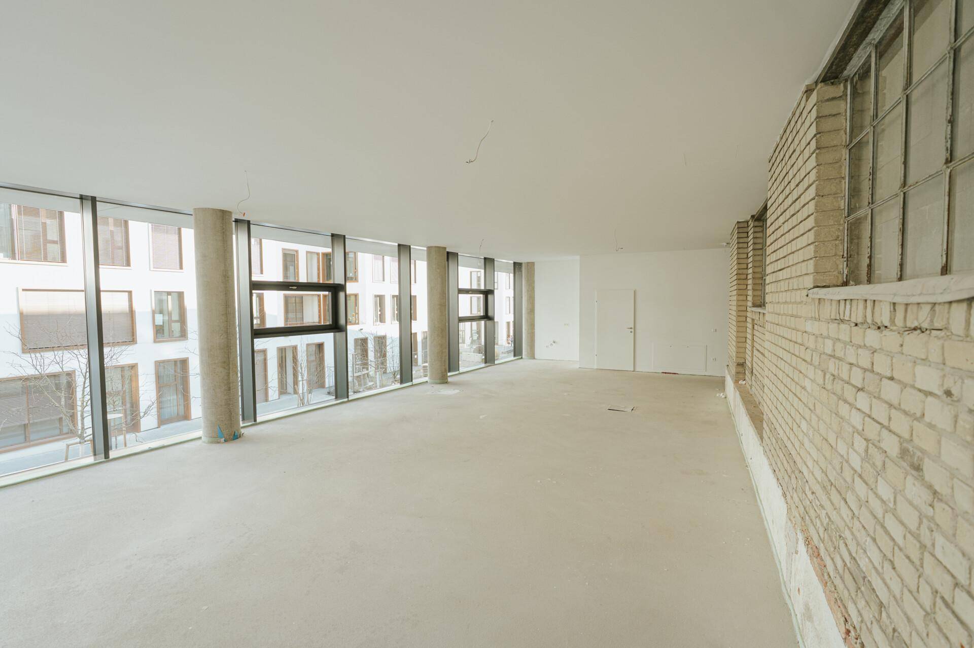 Großer, heller Büroraum mit durchgehender Fensterfront, Sichtbetonstützen und einer Ziegelwand.