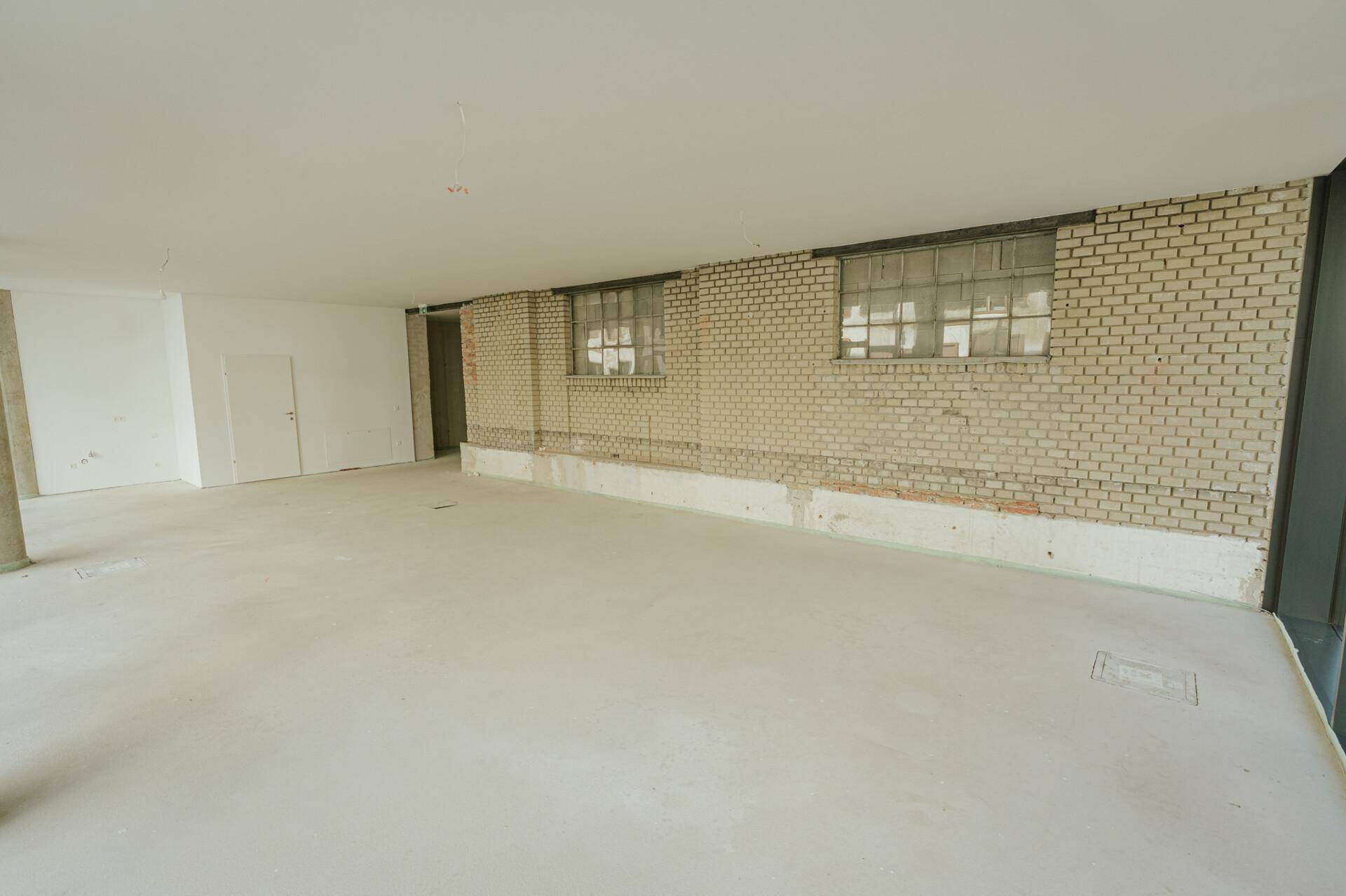 Geräumiger Gewerberaum mit unverputzter Ziegelwand, Betonboden und zwei Fenstern.
