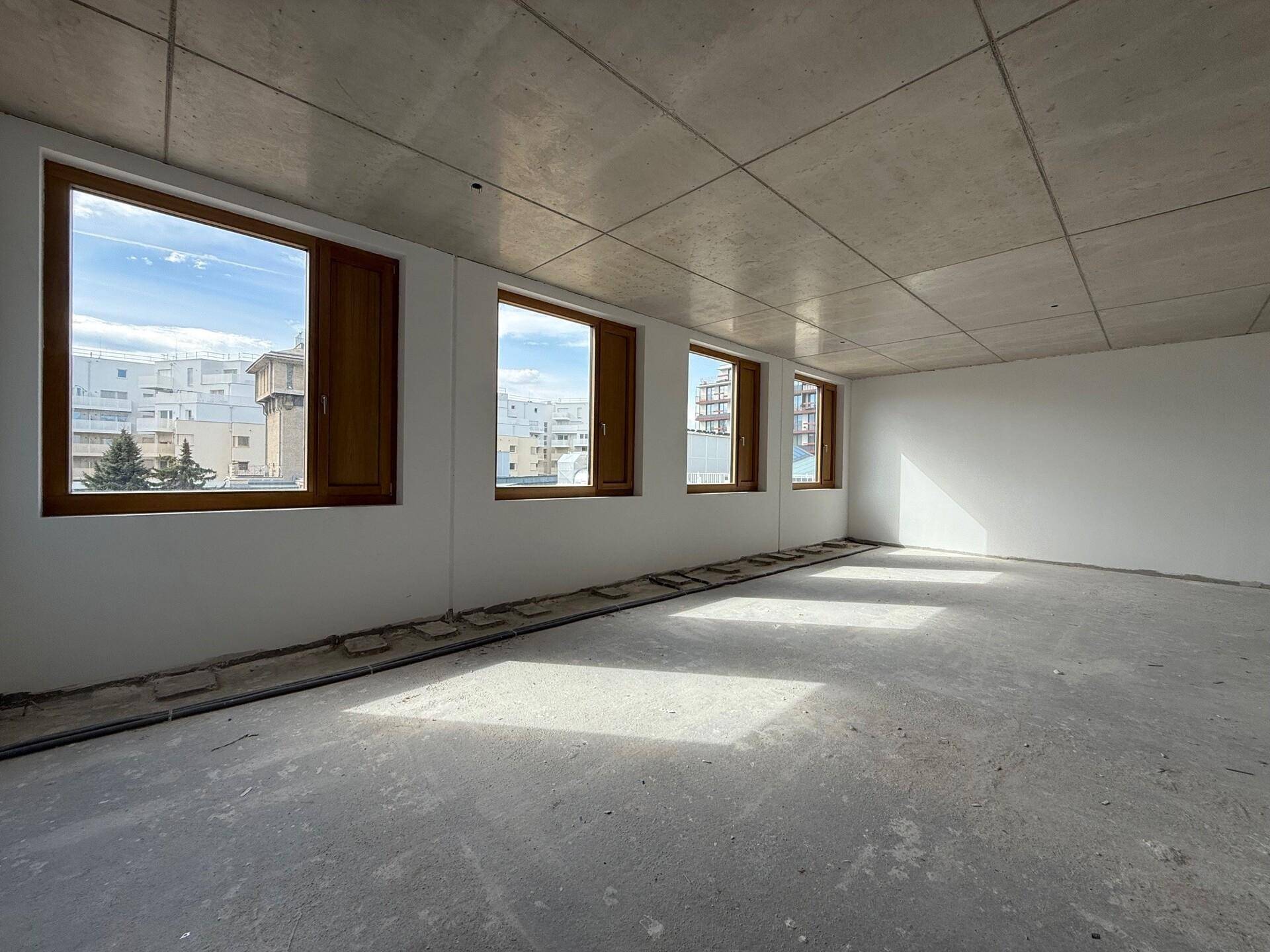Heller, leerer Raum mit weißen Wänden, Betondecke und Holzfenstern mit Stadtblick.