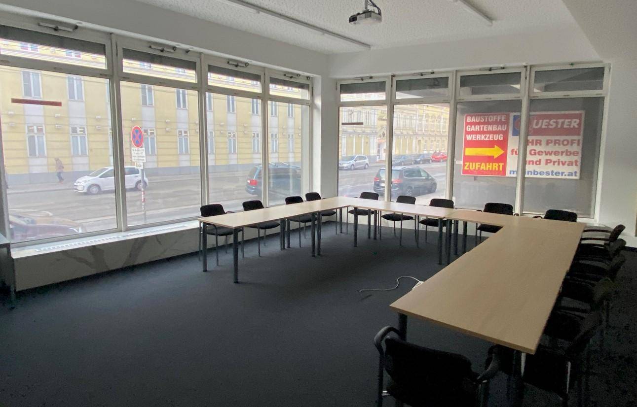 Konferenzraum mit Tischen und Stühlen, großen Fenstern zur Straße und viel Tageslicht.
