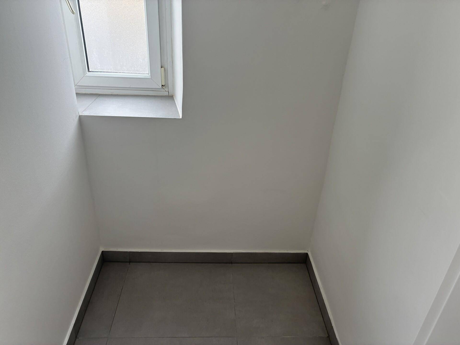 Kleiner Abstellraum mit Fenster, grauen Bodenfliesen und weißen Wänden.