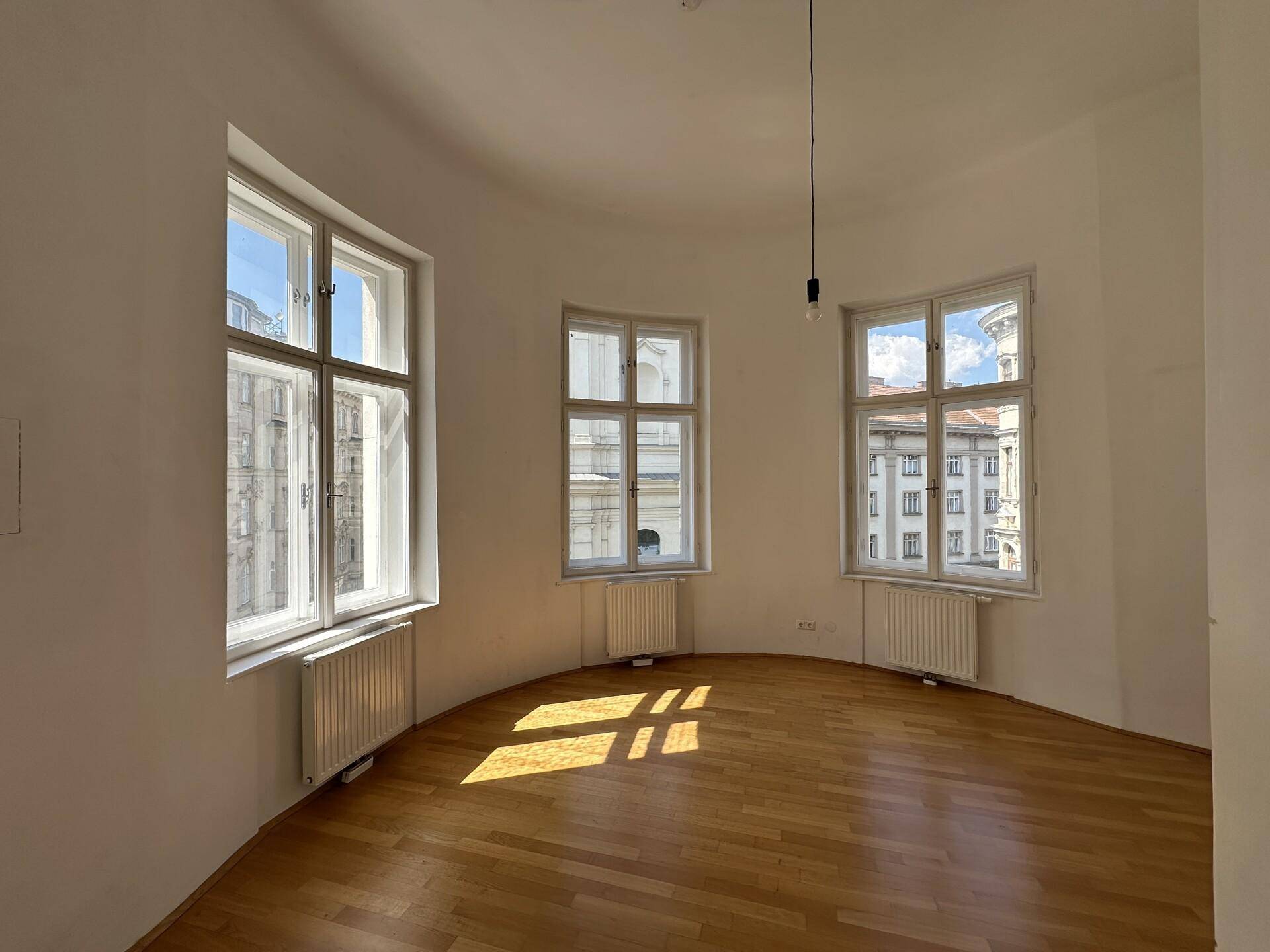 Helles, rundes Zimmer mit Parkettboden und mehreren Fenstern, das viel Tageslicht hereinlässt.