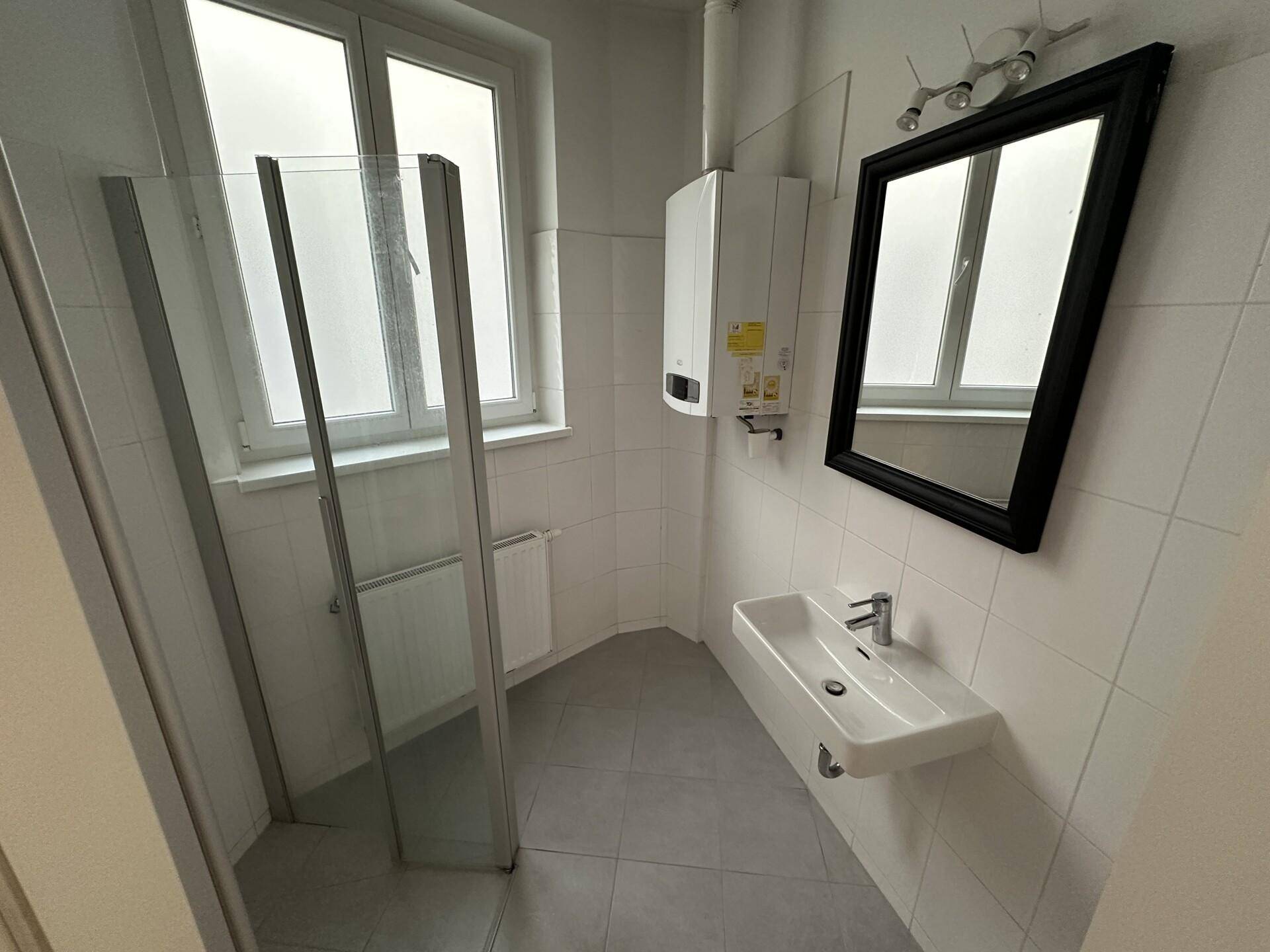 Badezimmer mit Dusche, Waschbecken und Fenster, ausgestattet mit weißen Fliesen und einem Boiler.