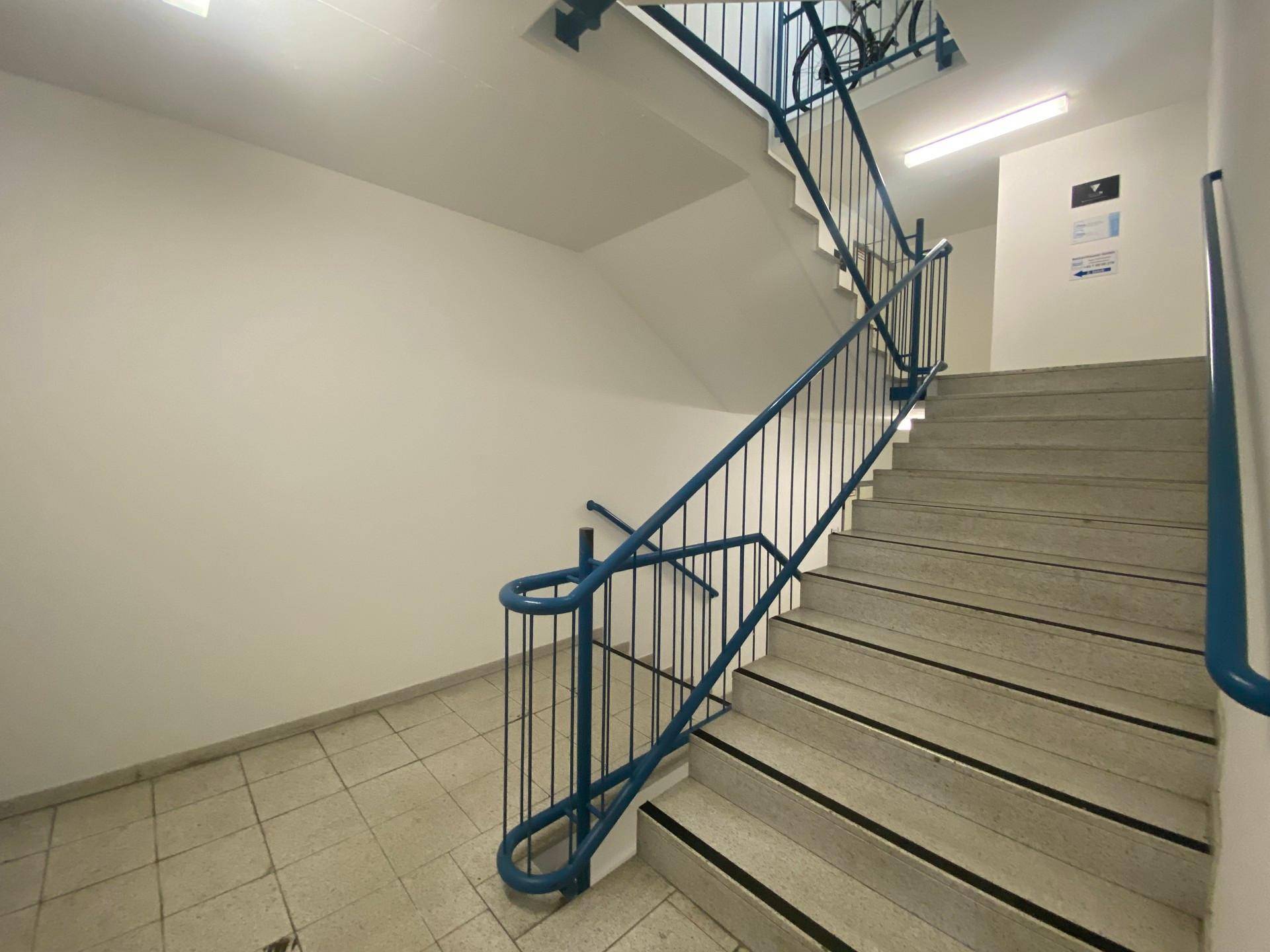 Treppenhaus mit gefliestem Boden, blauen Geländern und weißen Wänden, gut beleuchtet.