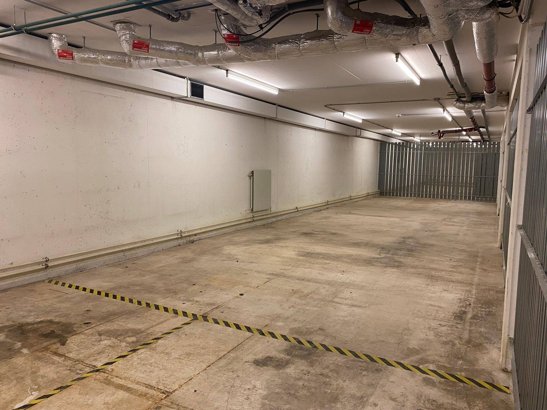 Langer Gang in der Tiefgarage mit Betonboden, gelb-schwarzen Markierungen und Lagerabteilen.