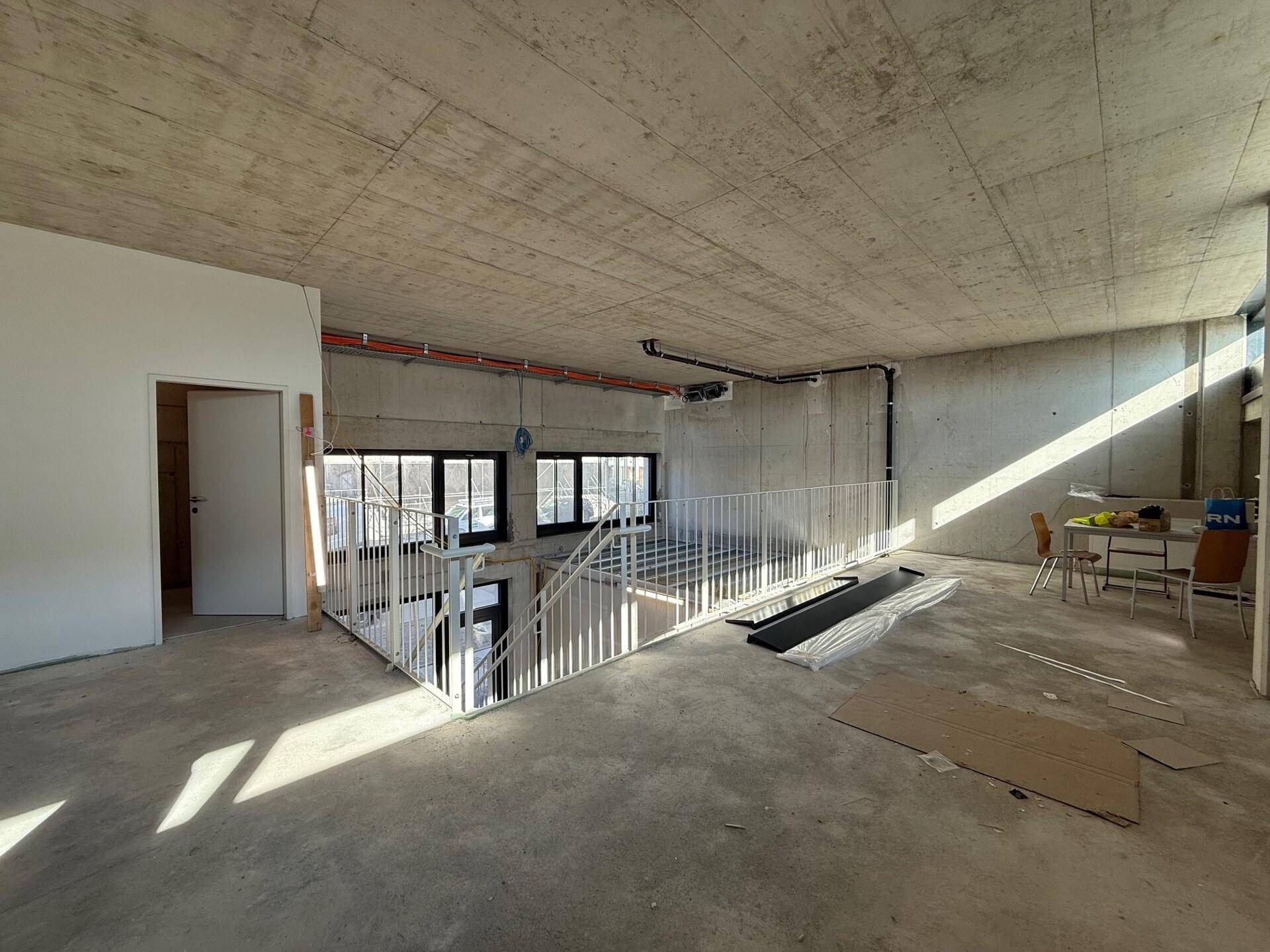 Großer offener Raum im Rohbau mit Sichtbetondecke und -boden, durchflutet von natürlichem Licht durch die Fenster.