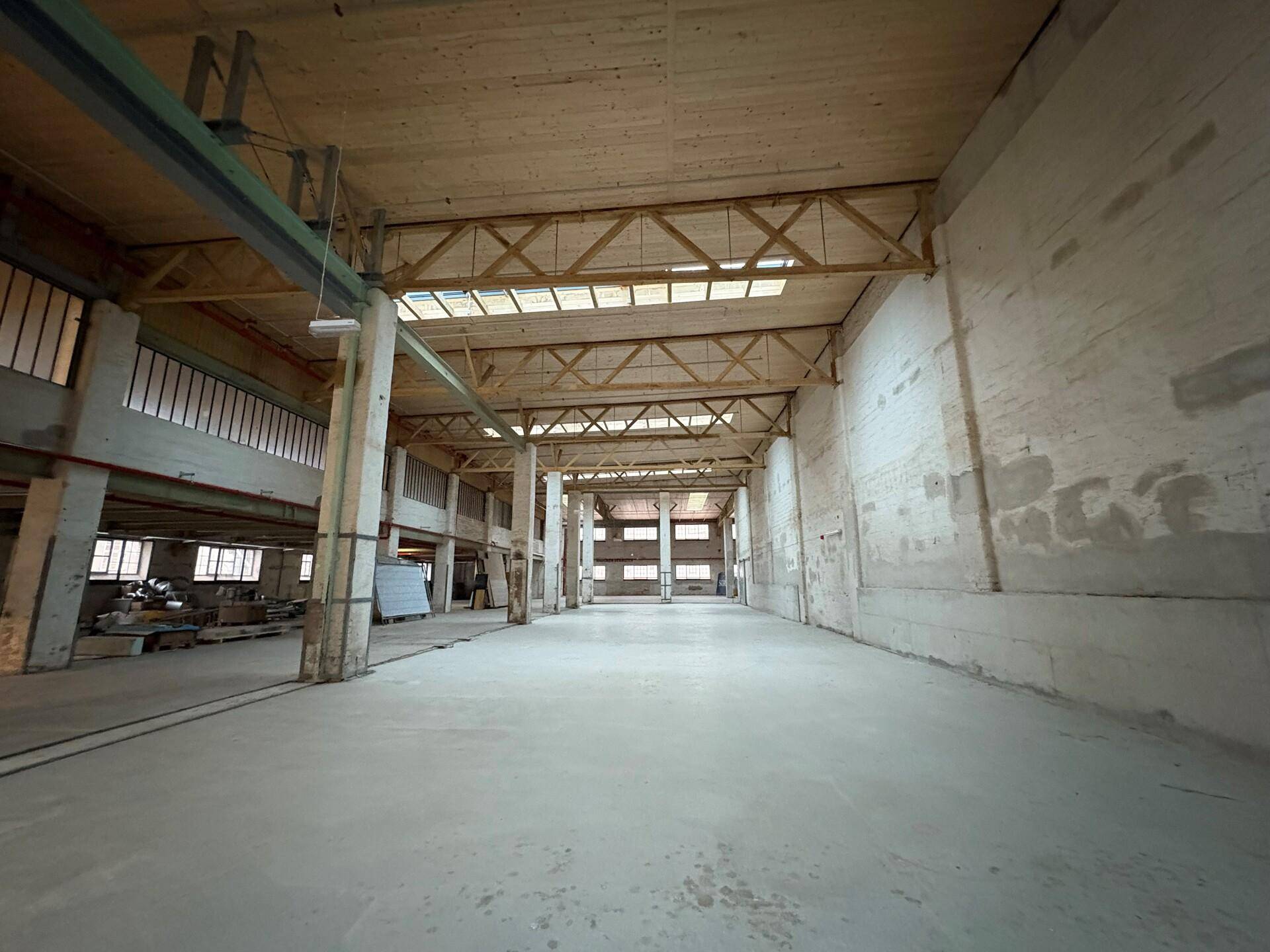 Weitläufige Lagerhalle mit hohen Decken, sichtbaren Holzbalken und großen Fenstern, die viel Tageslicht hereinlassen.