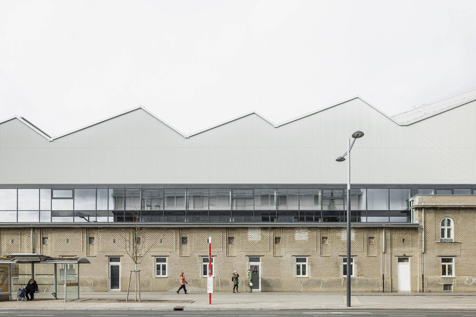 Eine lange Fassade mit Ziegelmauerwerk im unteren Bereich und einer modernen, gezackten Dachlinie darüber.