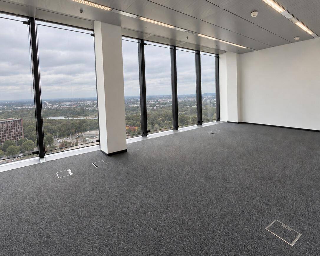 Leerer Büroraum mit großen Fenstern, grauem Teppichboden und weitem Blick über die Stadtlandschaft.