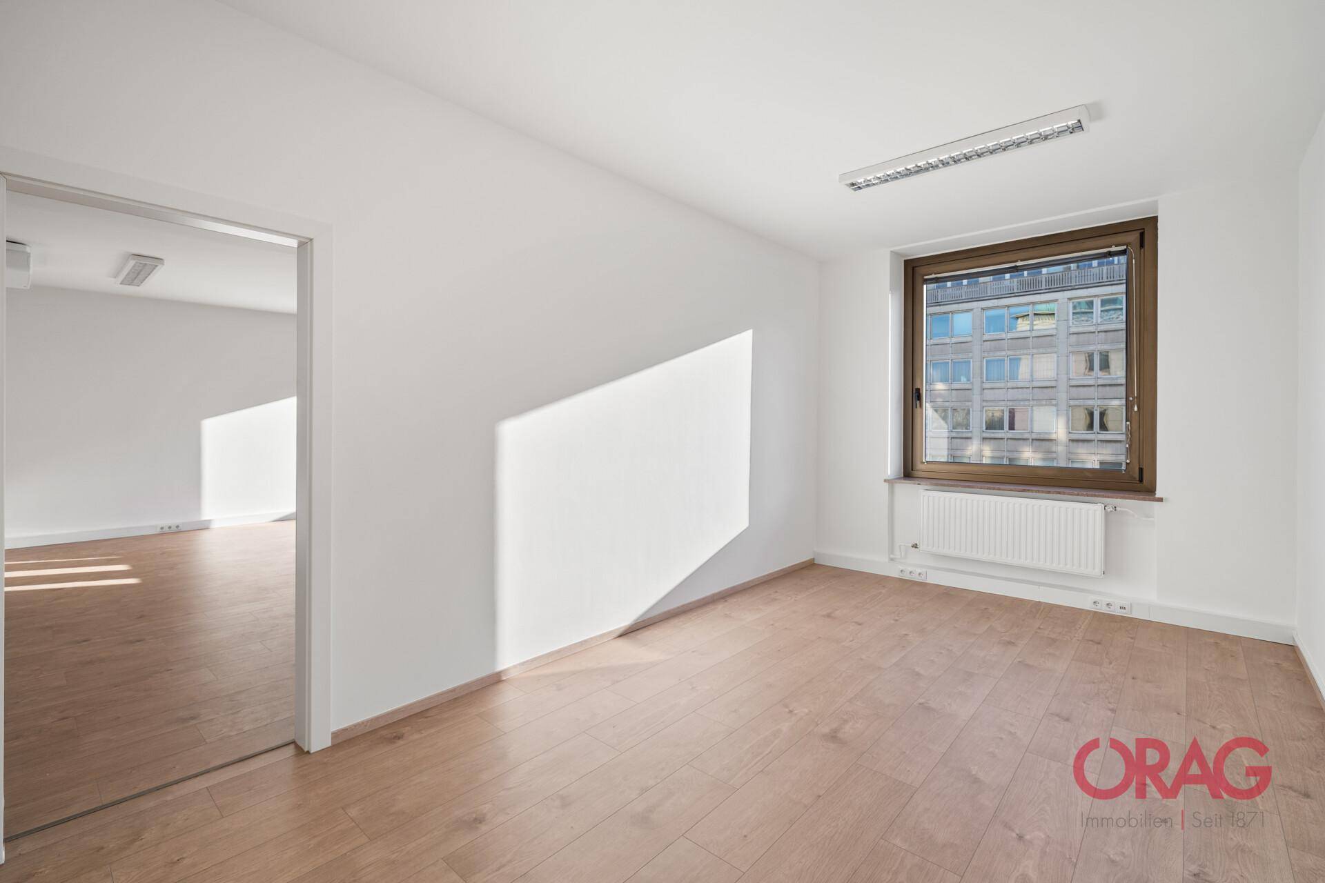 Leerer Büroraum mit hellem Holzboden, weißen Wänden und Blick auf die umliegenden Gebäude.