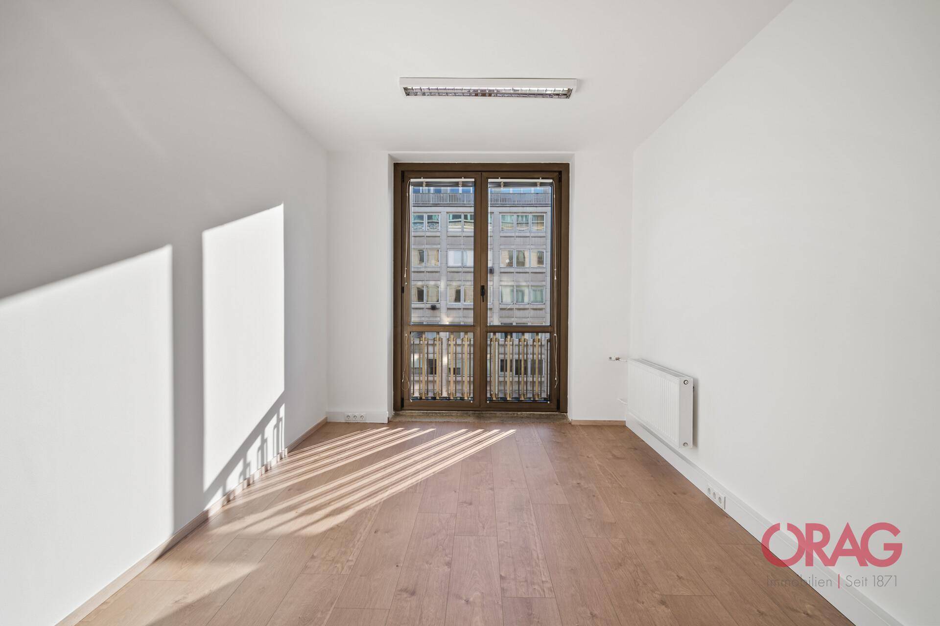 Heller Büroraum mit Holzboden, weißen Wänden und einer großen Fenstertür mit Stadtblick.