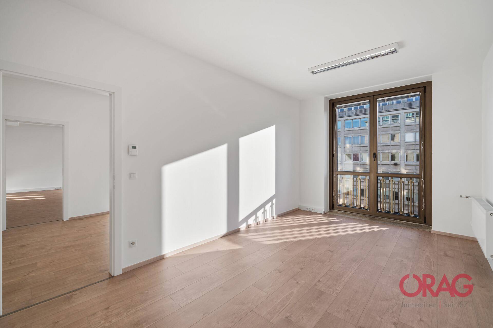 Heller Büroraum mit Holzboden, weißen Wänden und einer Fenstertür mit Blick auf die Stadt.