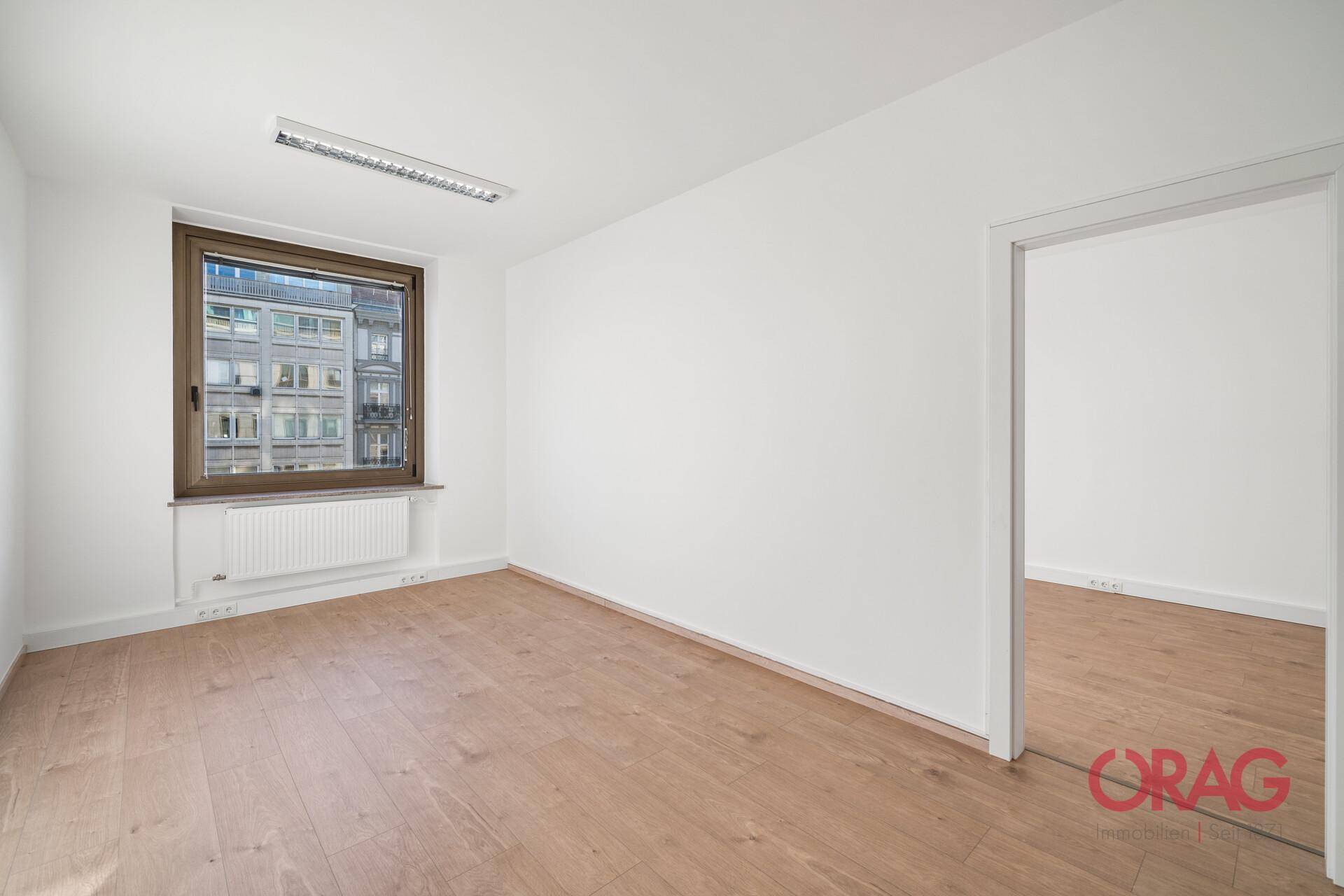 Leerer Büroraum mit Holzboden, weißen Wänden und einem Fenster mit Blick auf die Stadt.