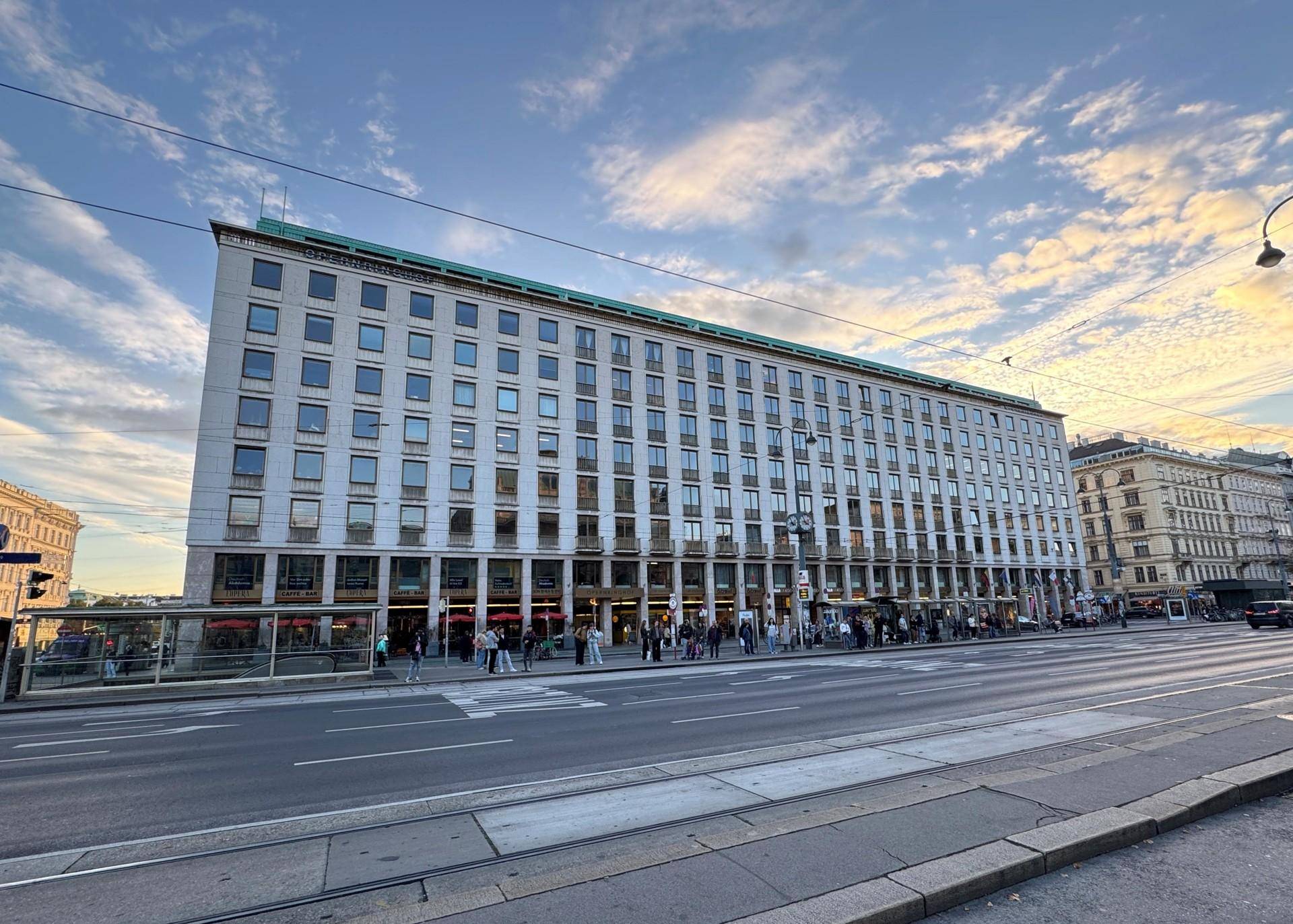 Gebäudefassade bei Sonnenuntergang mit beleuchteten Fenstern und belebter Straße davor.