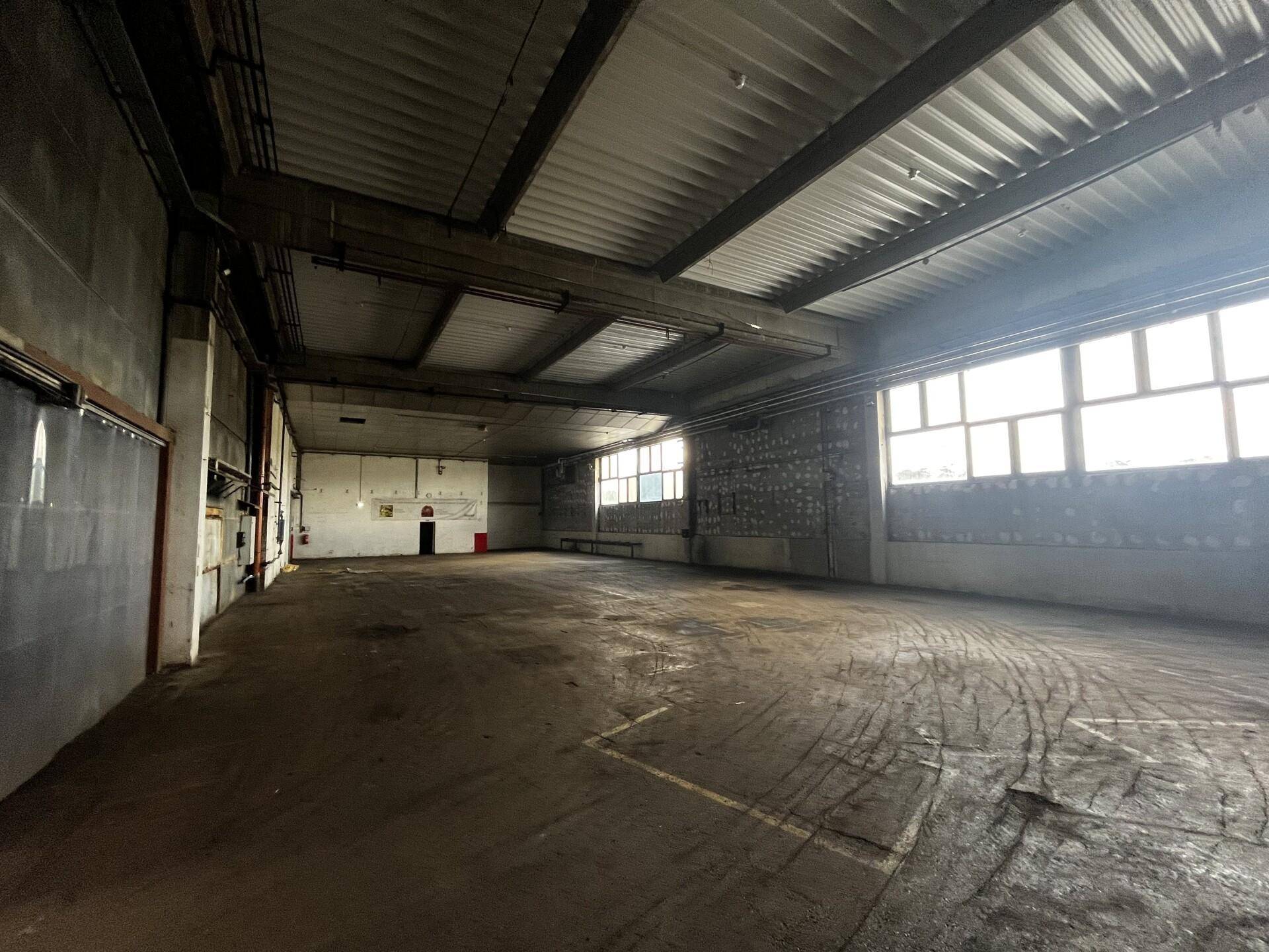 Leerer Industrieraum mit Betonboden, Wellblechdecke und großen Fenstern, ideal für Werkstatt oder Lager.