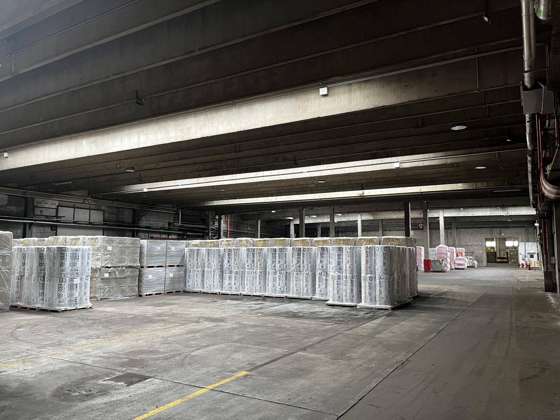Lagerhalle mit gestapelten Waren auf Paletten, Betonbalken und Beleuchtung.