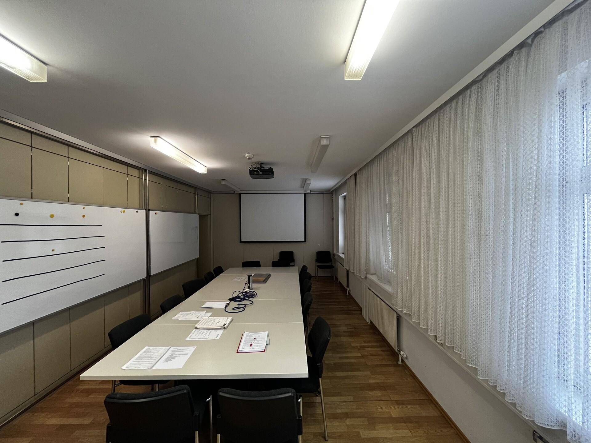 Konferenzraum mit langem Tisch, Stühlen, Projektionsleinwand und Whiteboards, ideal für Besprechungen.