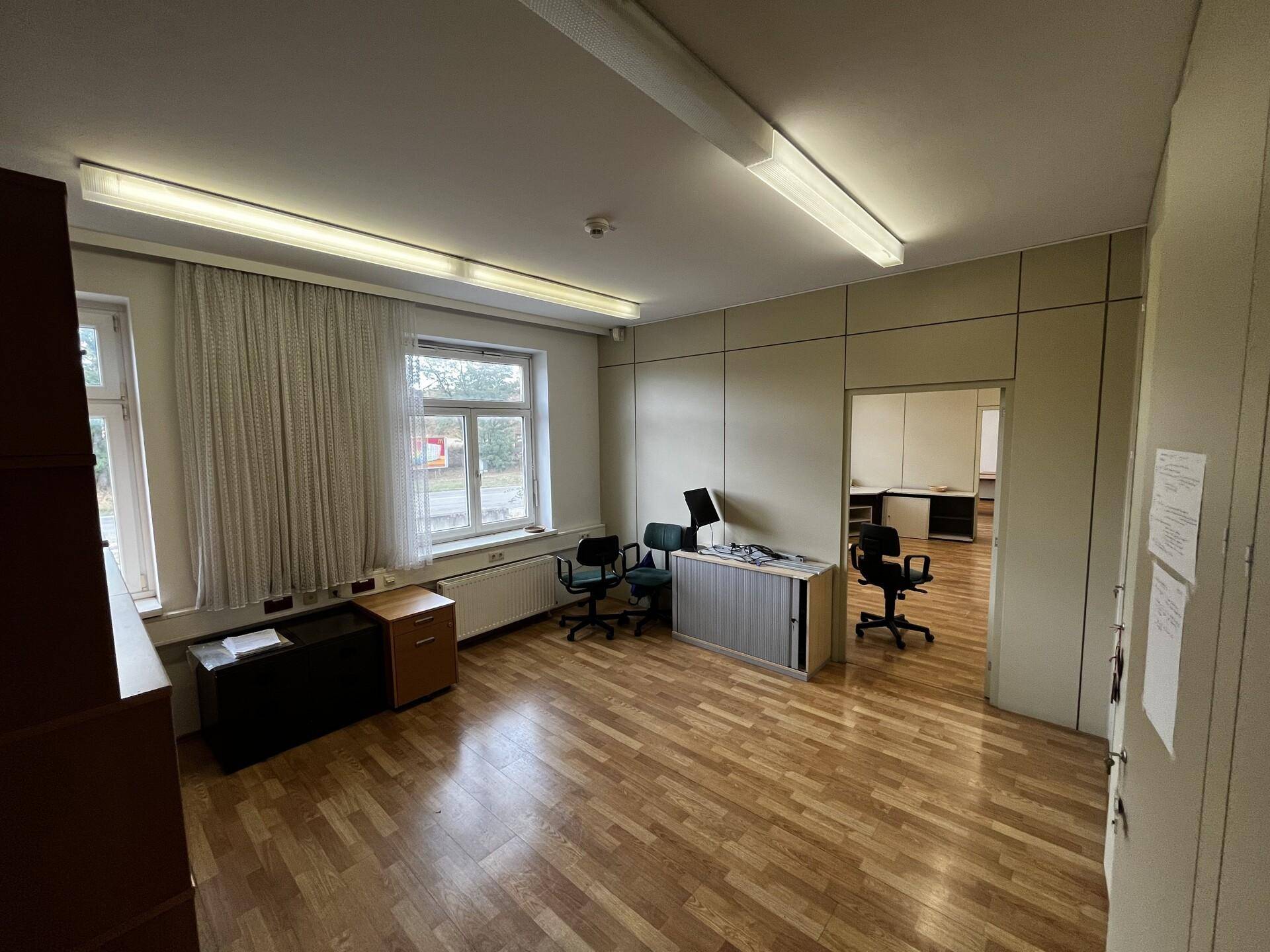 Büroraum mit Holzparkettboden, großem Fenster und Büromöbeln, angrenzend an weiteres Büro.