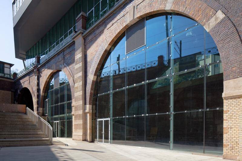 Historische Ziegelbögen mit großen, modernen Glasfronten, die einen Eingangsbereich bilden, daneben eine Treppe.