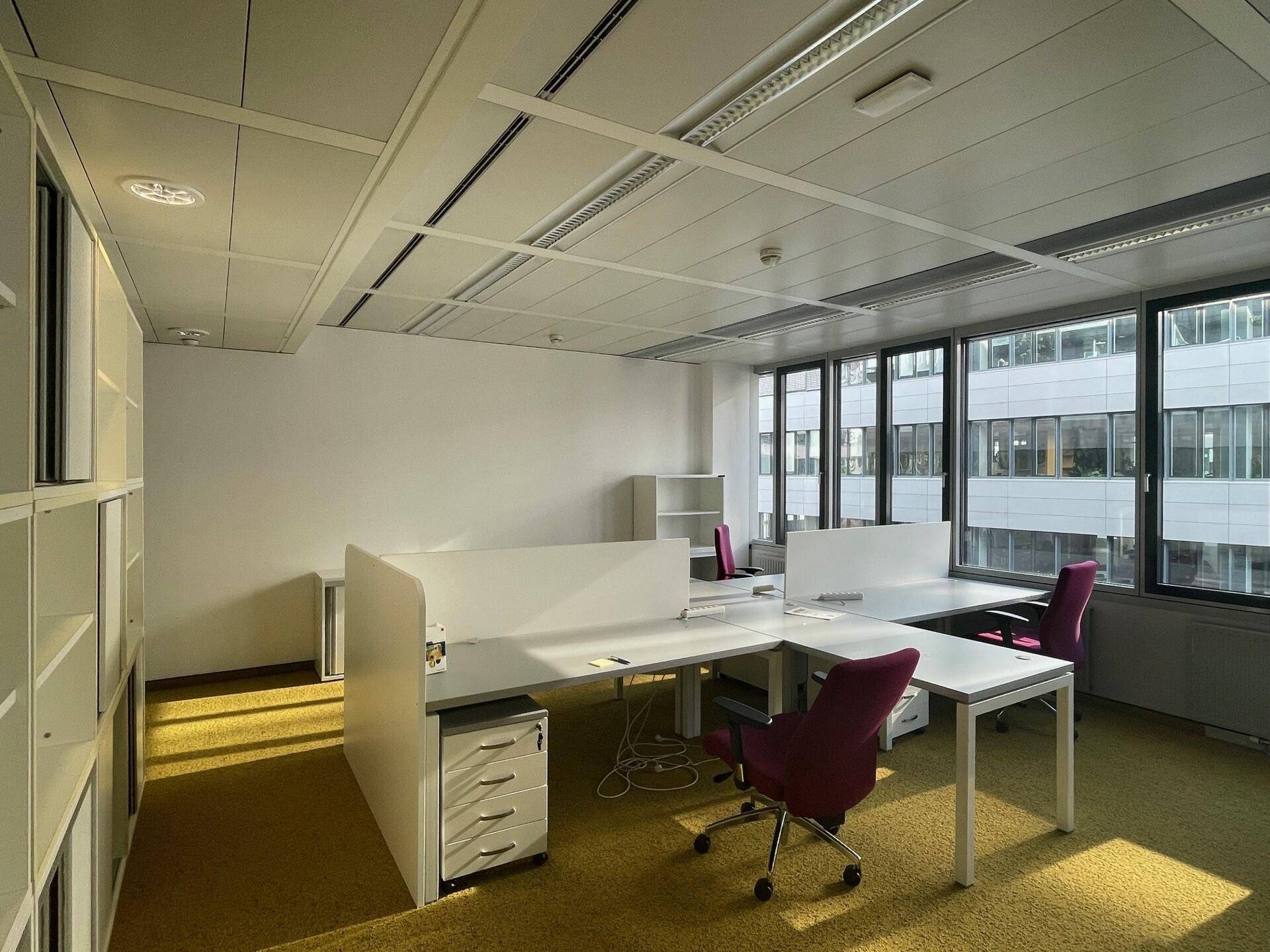 Heller Büroraum mit mehreren Arbeitsplätzen, gelbem Teppichboden und großen Fenstern mit Stadtblick.