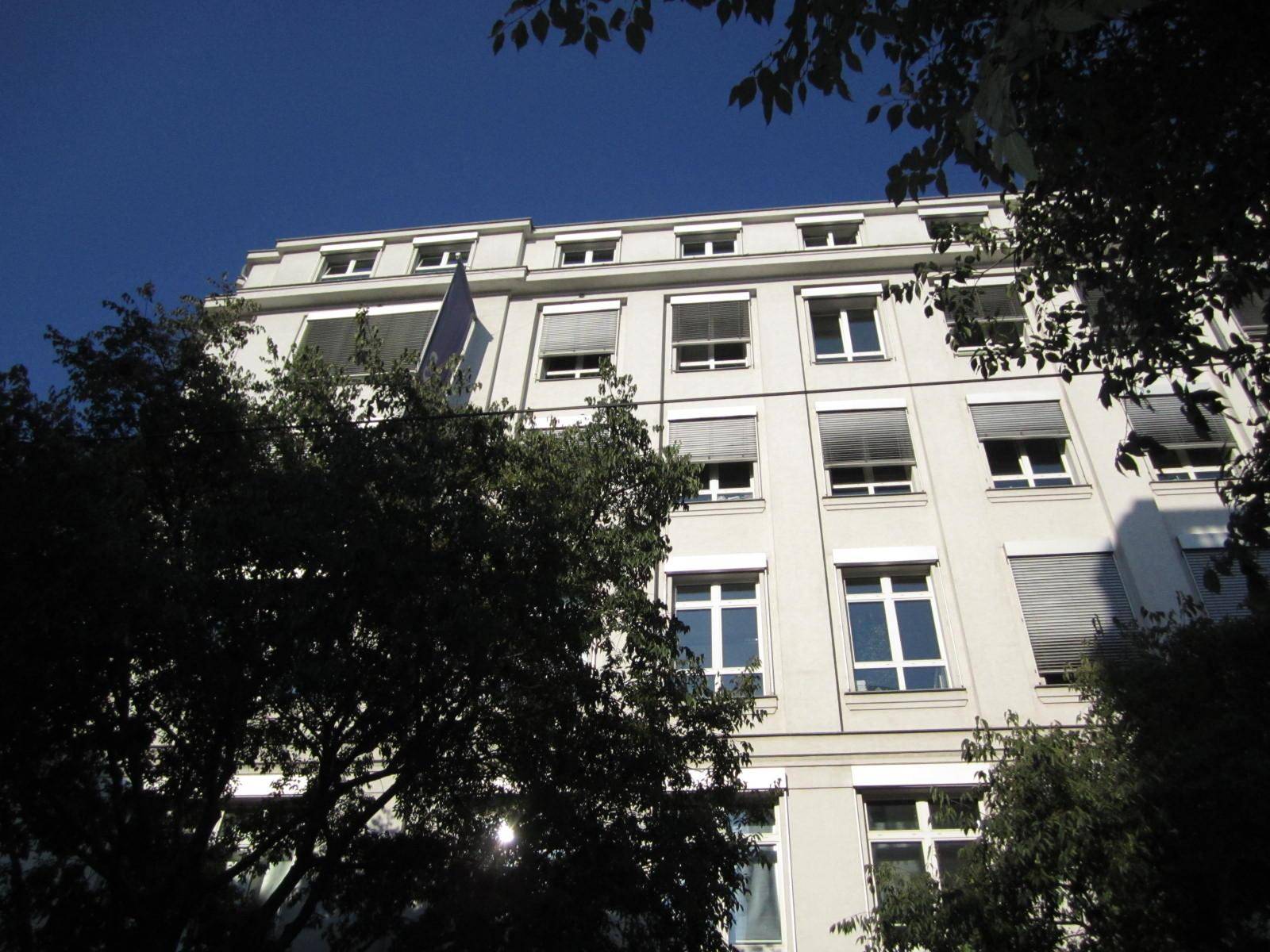 Weiße Gebäudefassade mit vielen Fenstern und Jalousien, umgeben von Bäumen unter blauem Himmel.