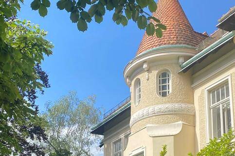Historisches Gebäude mit markantem Turm, runden Fenstern und grüner Umgebung unter blauem Himmel.