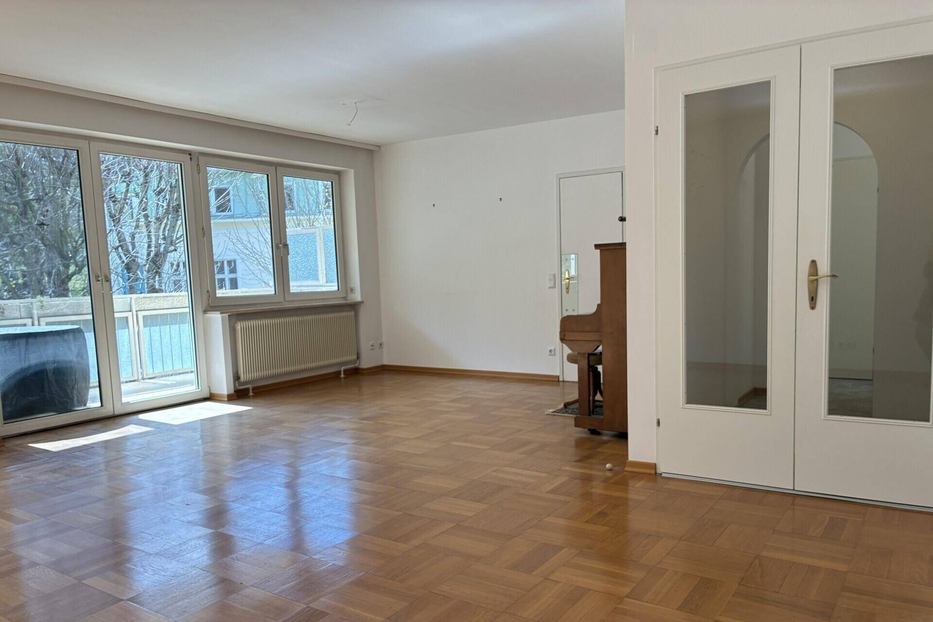 Geräumiges Wohnzimmer mit Parkettboden, großen Fenstern und einem Klavier im Hintergrund.