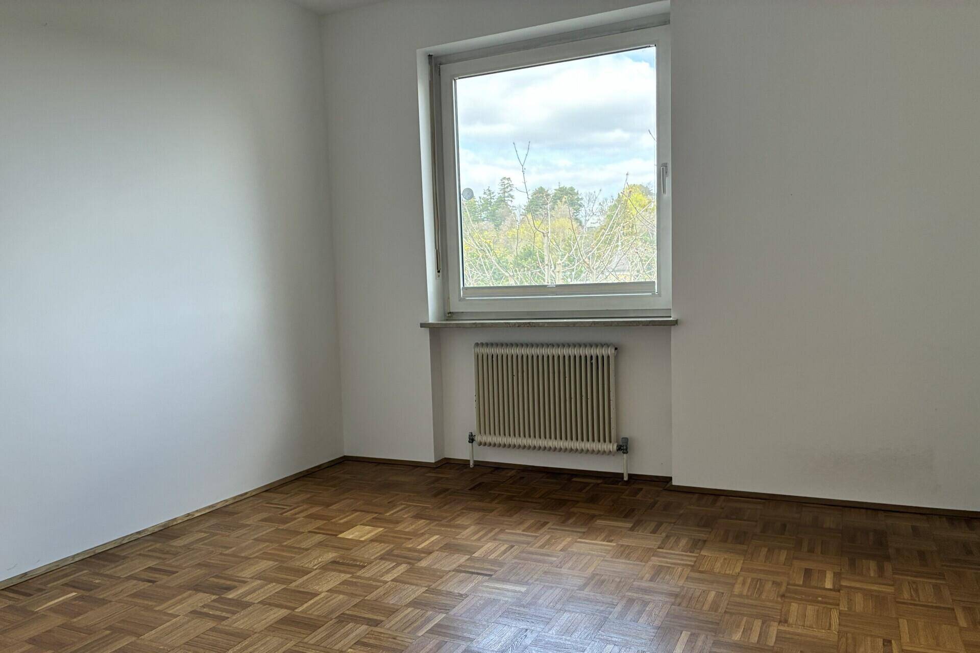 Leeres Schlafzimmer mit Parkettboden und großem Fenster, das viel Tageslicht hereinlässt.