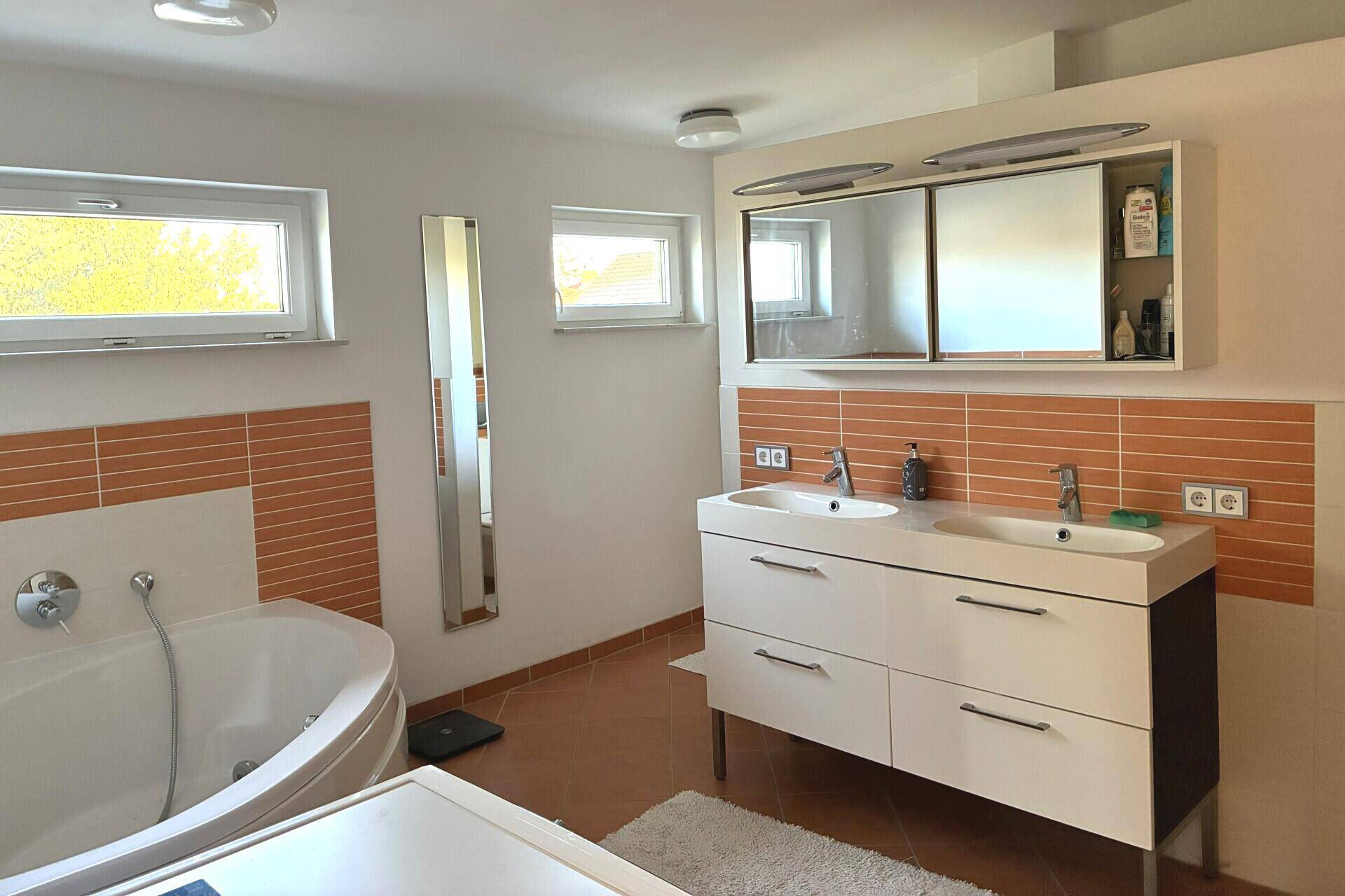 Helles Badezimmer mit Doppelwaschbecken, Eckbadewanne und Fenster.