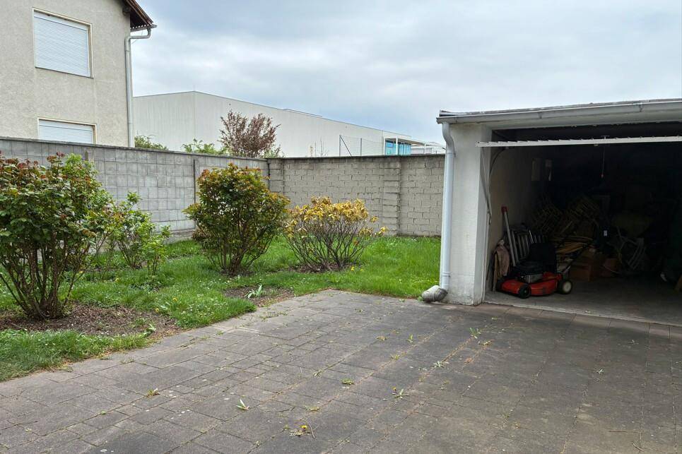 Offene Garage mit Geräten und ein gepflasterter Bereich neben einem kleinen Gartenabschnitt.