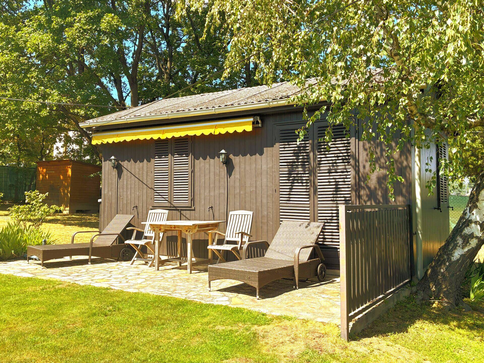 Sonnige Terrasse mit Liegestühlen und einem Tisch vor einem kleinen Holzhaus mit gelber Markise.