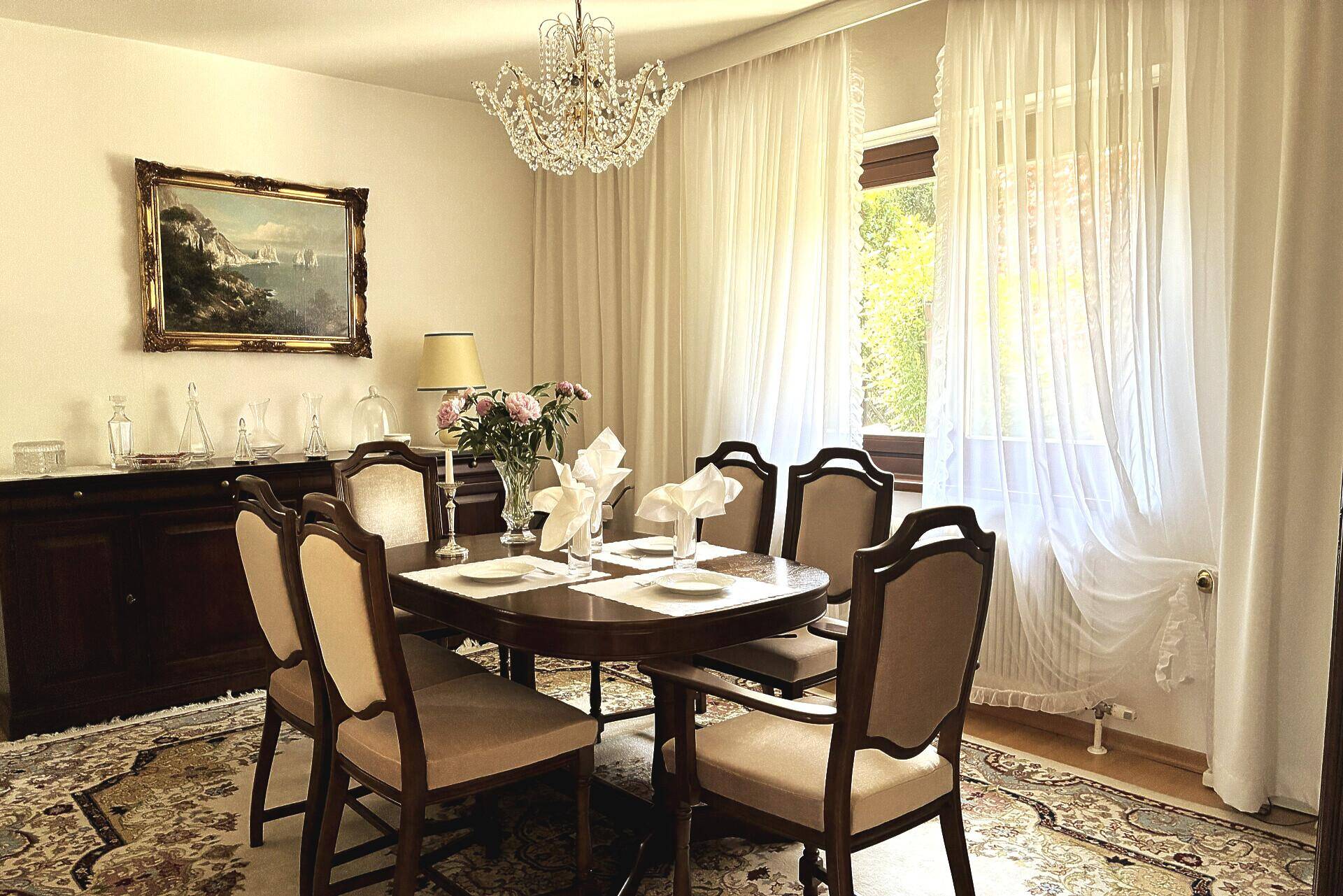 Eleganter Essbereich mit klassischem Holztisch, Kronleuchter und großen Fenstern mit Vorhängen.