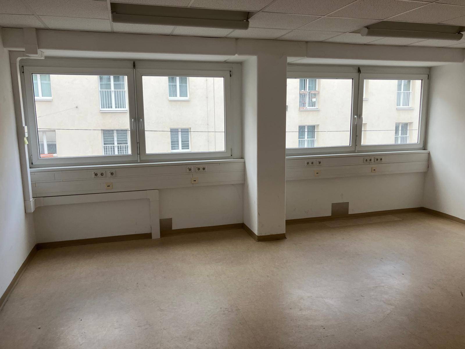 Geräumiges, leeres Büro mit mehreren Fenstern, die einen Blick auf die gegenüberliegende Bebauung bieten.