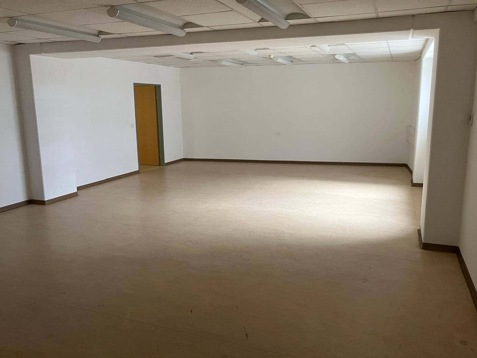 Großes, leeres Büro mit hellen Wänden, abgehängter Decke und einem Zugang zu einem weiteren Bereich.