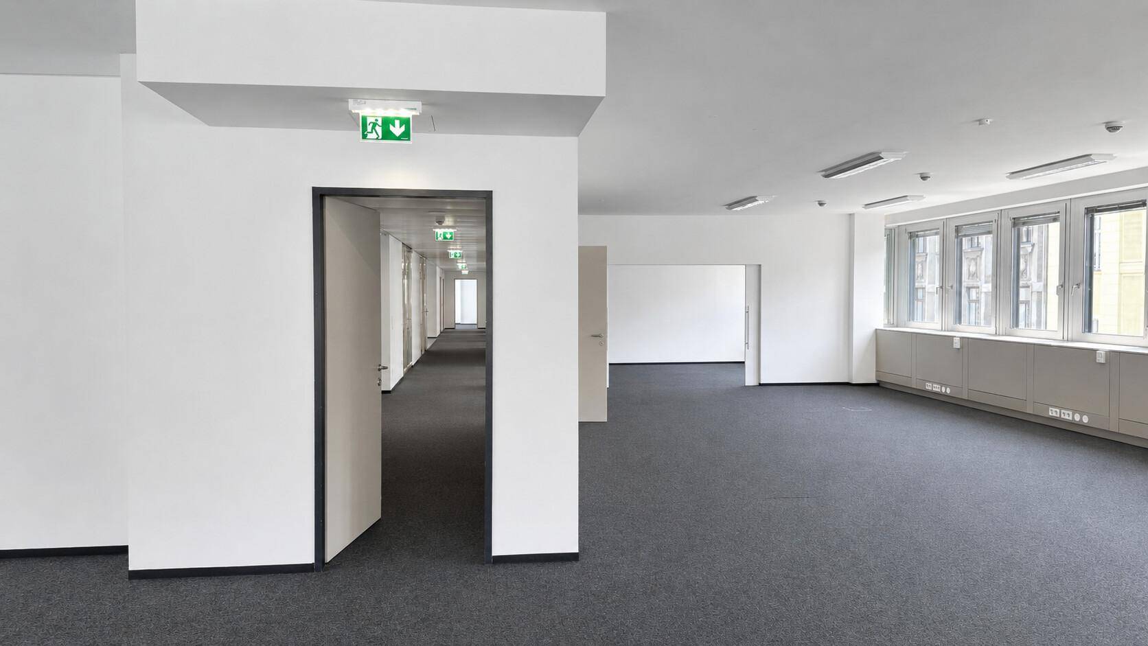 Geräumiges Großraumbüro mit Teppichboden, weißen Wänden und Zugang zu einem langen Flur.
