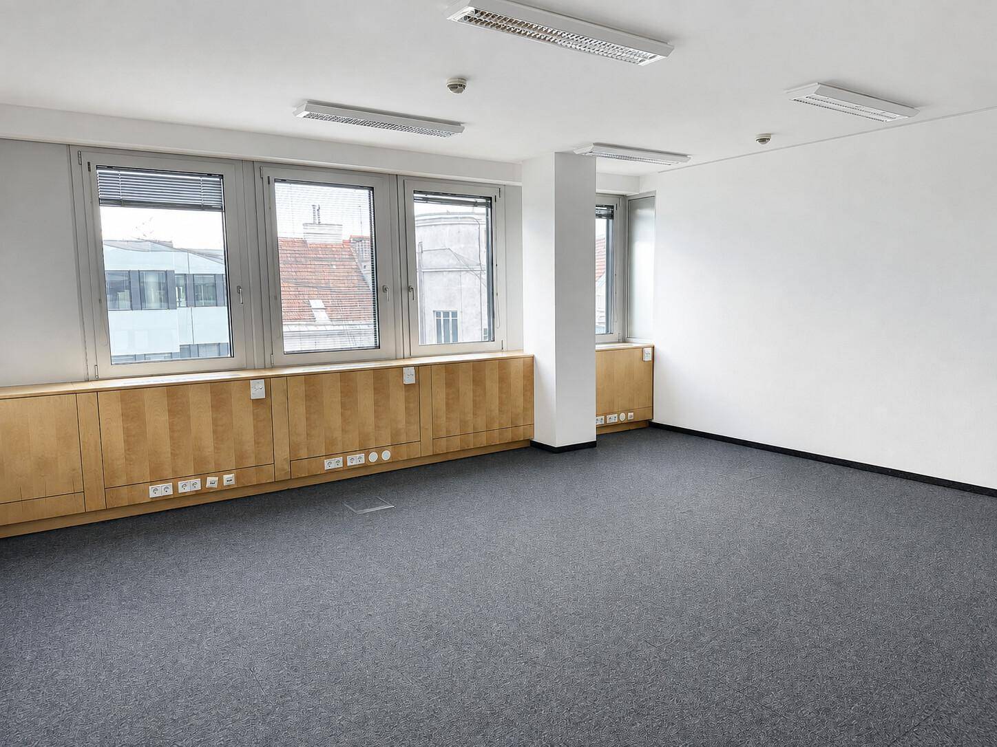 Leeres Büro mit Teppichboden, großen Fenstern und integrierten Holzschränken unter den Fenstern.