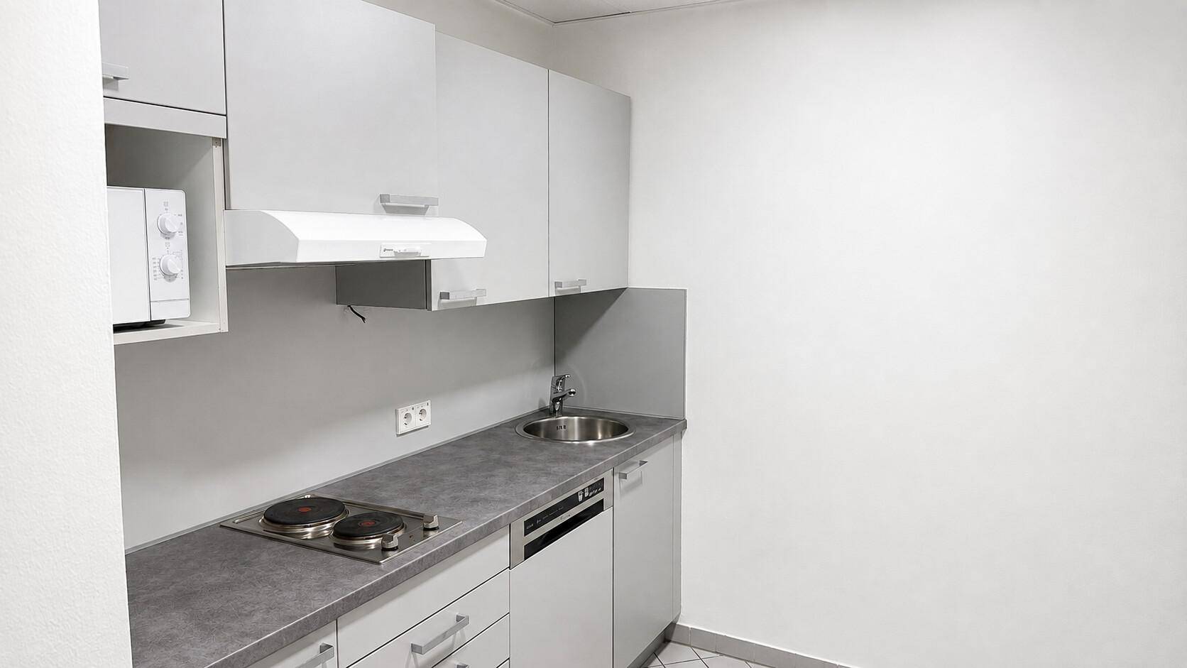 Kleine, funktionale Küchenzeile mit Spüle, Kochfeld, Mikrowelle und grauen Schränken.