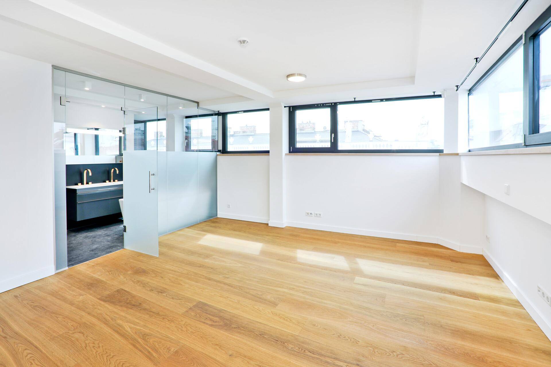 Helles Schlafzimmer mit Holzboden und angrenzendem, durch Glas abgetrenntem Badezimmer.