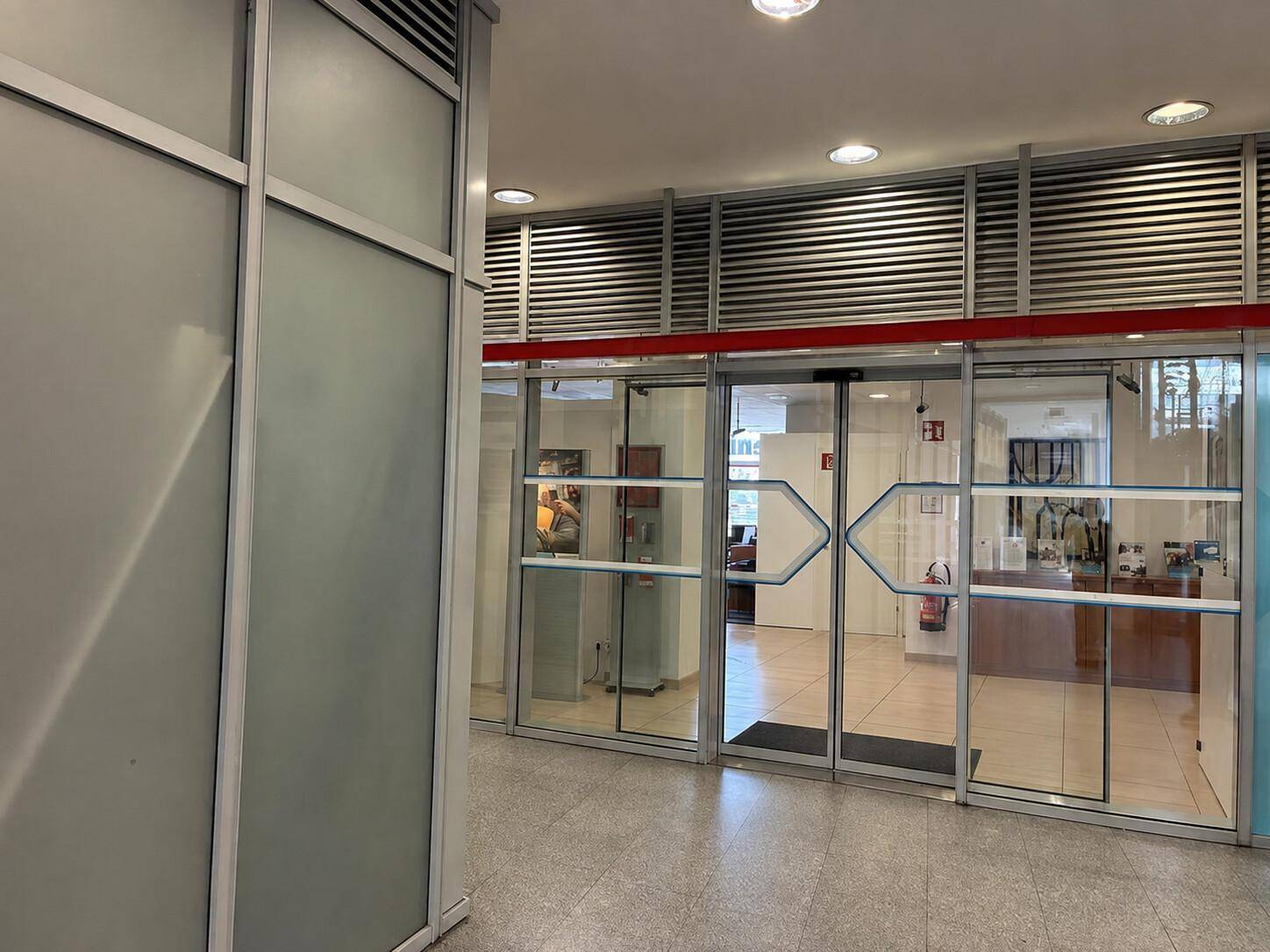 Eingangsbereich mit automatischen Glastüren, Fliesenboden und Deckenbeleuchtung.
