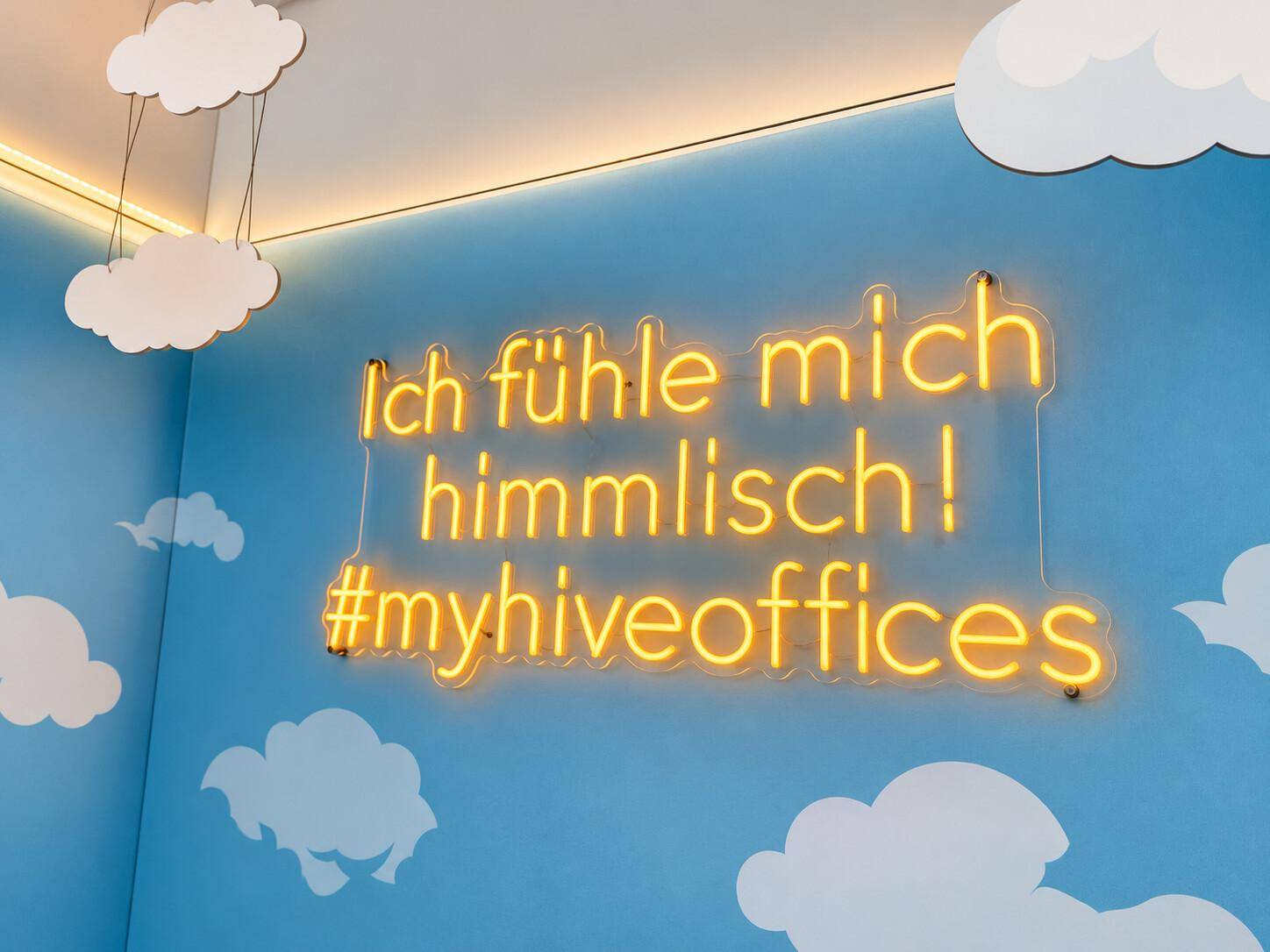 Eine blaue Wand mit Wolkenmotiven und einem leuchtenden Neonschild mit inspirierender Botschaft.