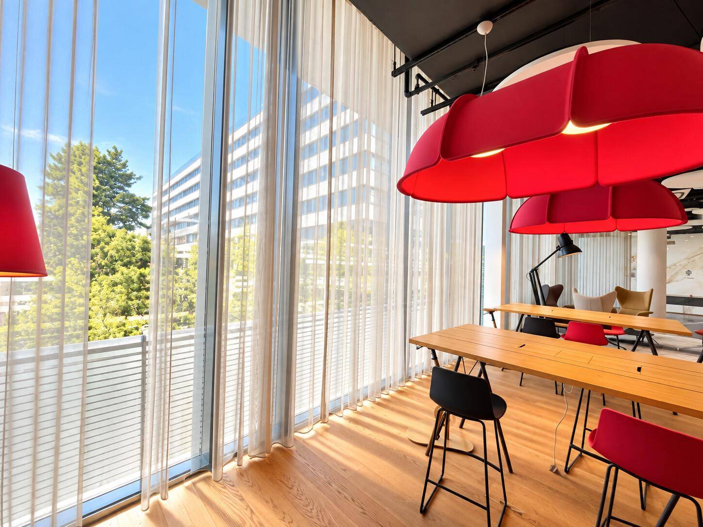 Heller Lounge-Bereich mit Holztischen, roten Designerlampen und großen Fenstern mit Blick nach draußen.