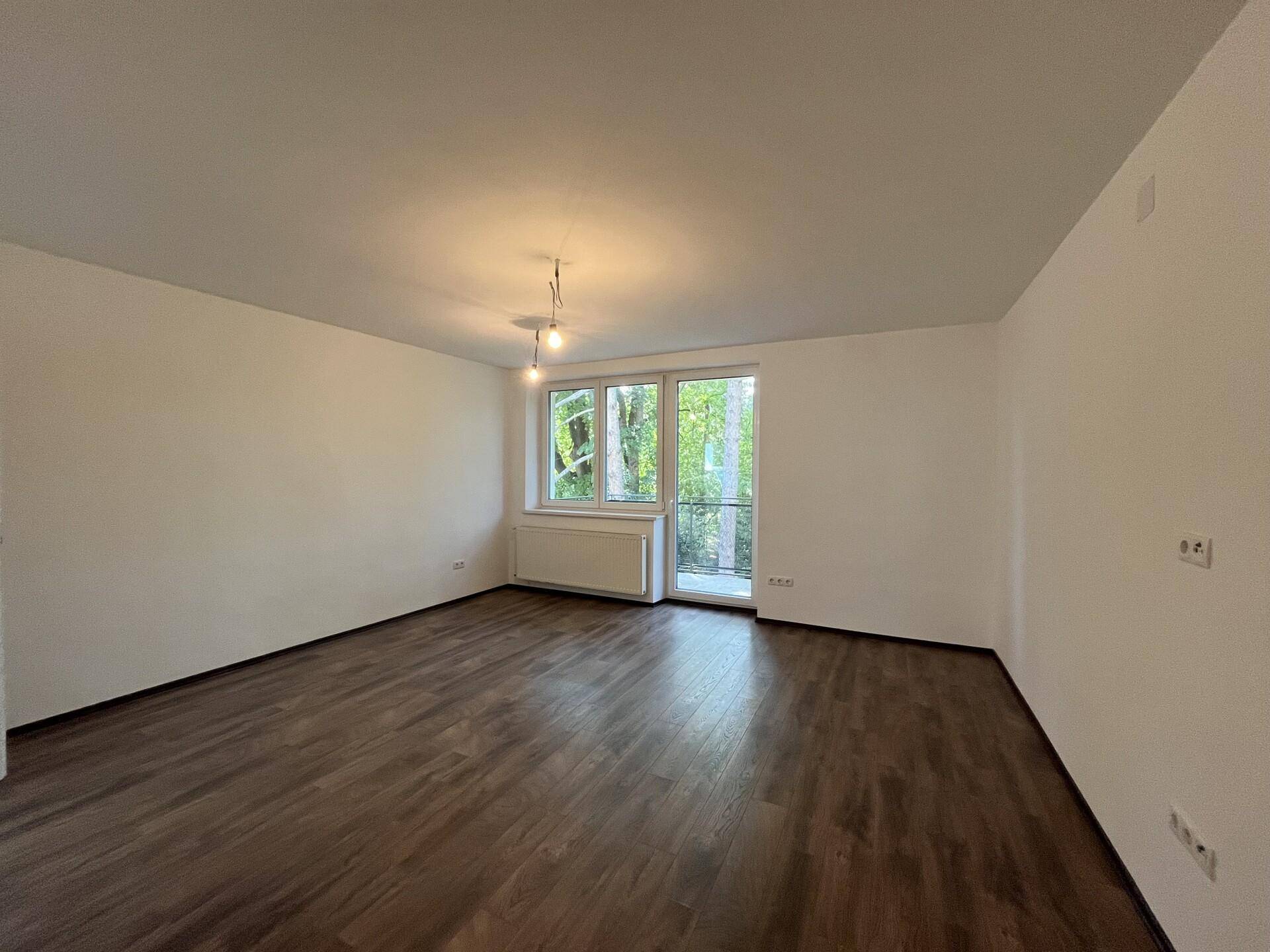 Heller, leerer Wohnraum mit dunklem Laminatboden und Fensterfront zum Balkon.