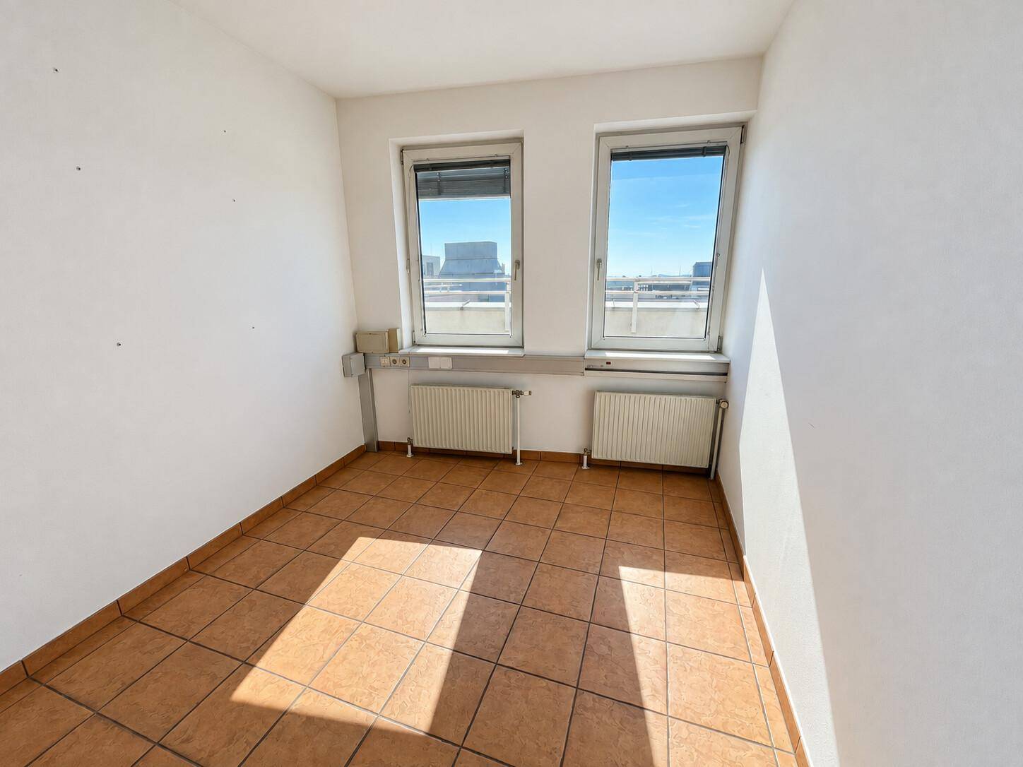 Kleinerer Büroraum mit Fliesenboden und zwei Fenstern, die viel Tageslicht hereinlassen.