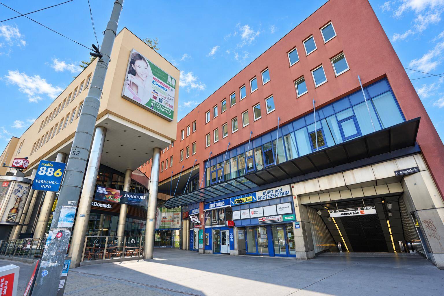Modernes Geschäftsgebäude mit U-Bahn-Zugang und verschiedenen Geschäften im Erdgeschoss in urbaner Umgebung.