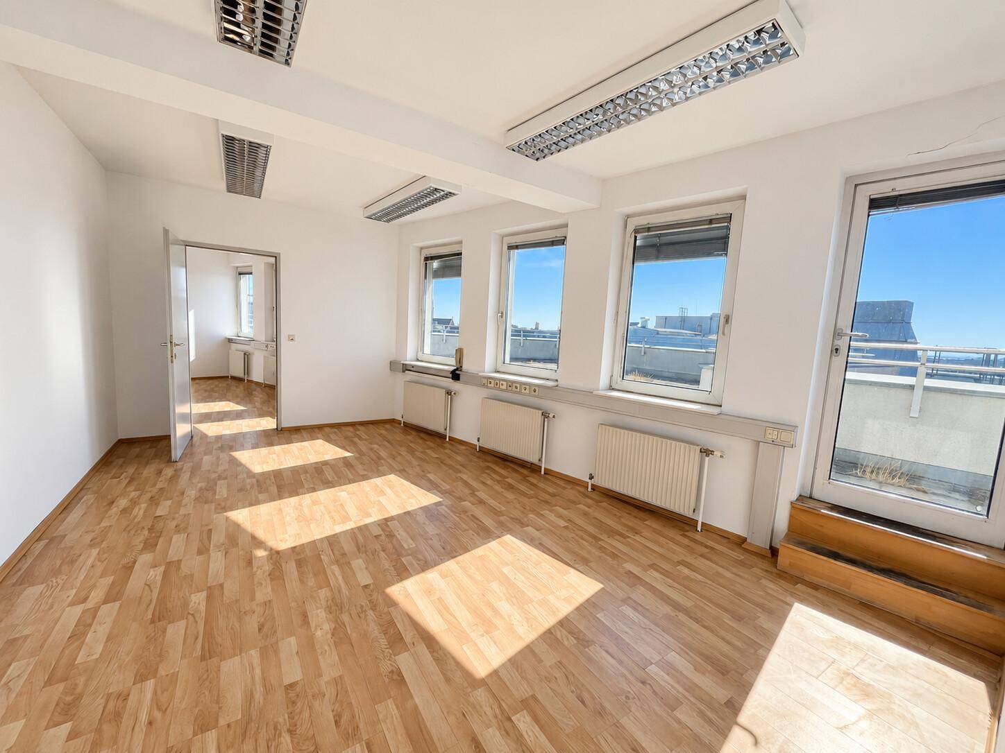 Geräumiger Büroraum mit Laminatboden, Deckenleuchten und großen Fenstern mit Balkonzugang.