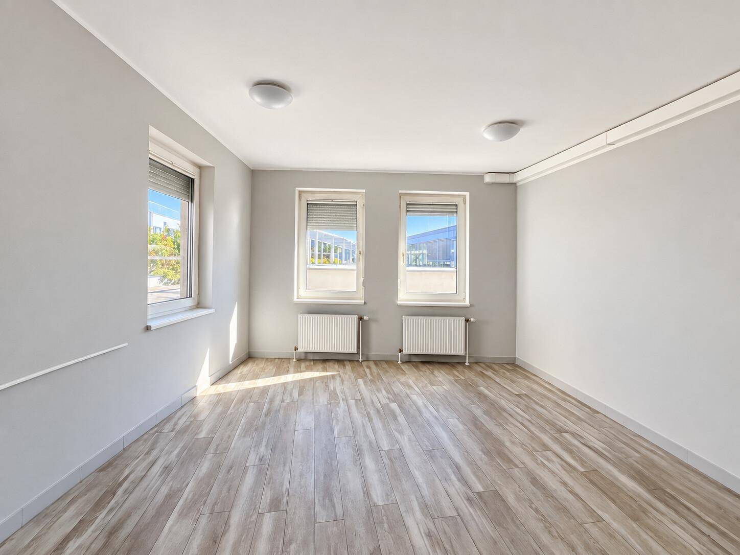 Großer, heller Büroraum mit mehreren Fenstern, die viel Tageslicht hereinlassen, und Holzoptikboden.