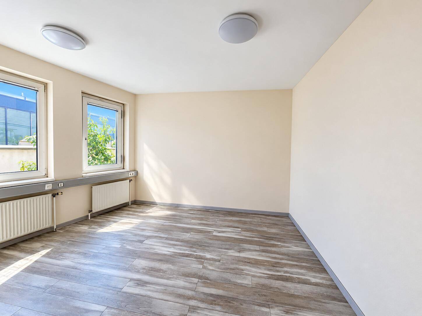 Geräumiger Büroraum mit hellen Wänden, Holzoptikboden und viel Tageslicht durch die großen Fenster.