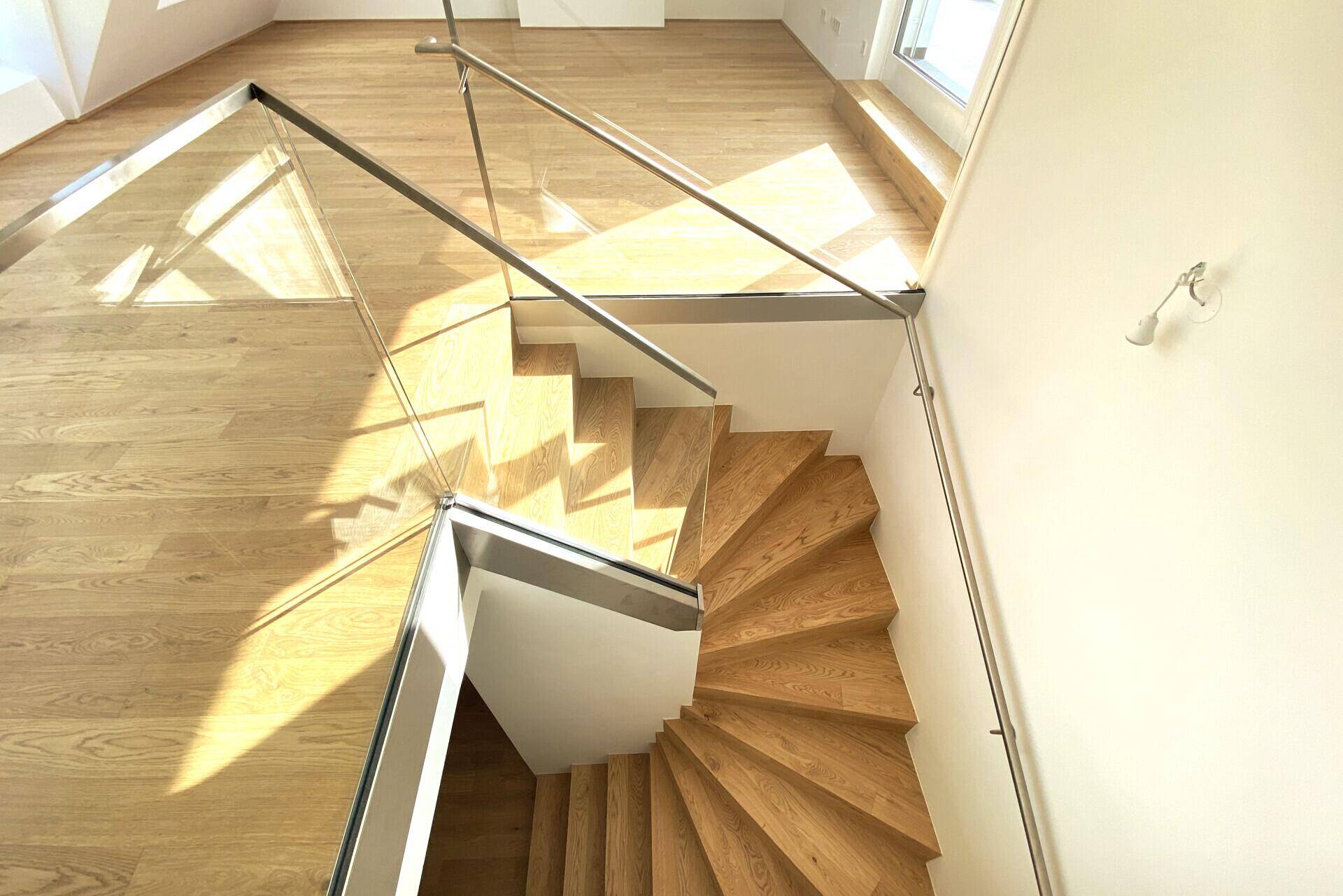 Moderne Holztreppe mit Glasgeländer, die zu einem hellen Wohnbereich im Dachgeschoss führt.