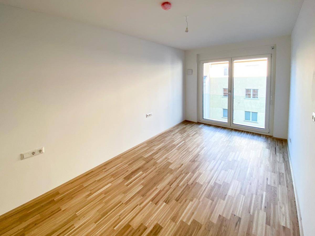Heller, leerer Raum mit Parkettboden und großem Fenster zur Terrasse oder Balkon.